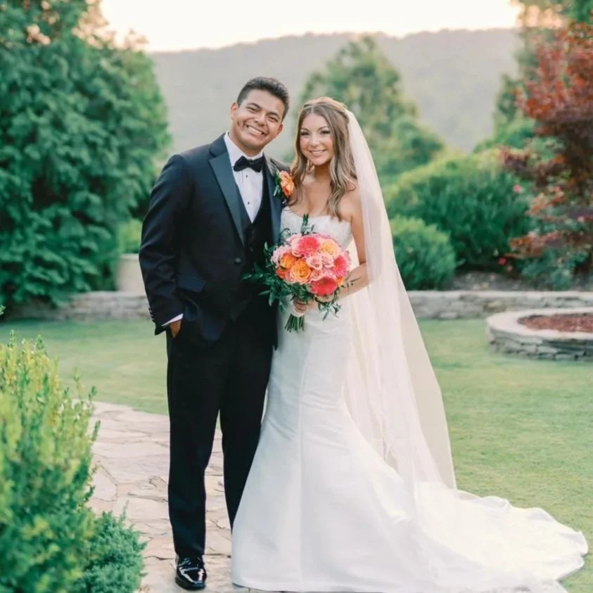 A smiling bride and groom standing outdoors on their wedding day, with the bride holding a bouquet of pink and orange flowers, and both dressed in formal wedding attire, surrounded by greenery.