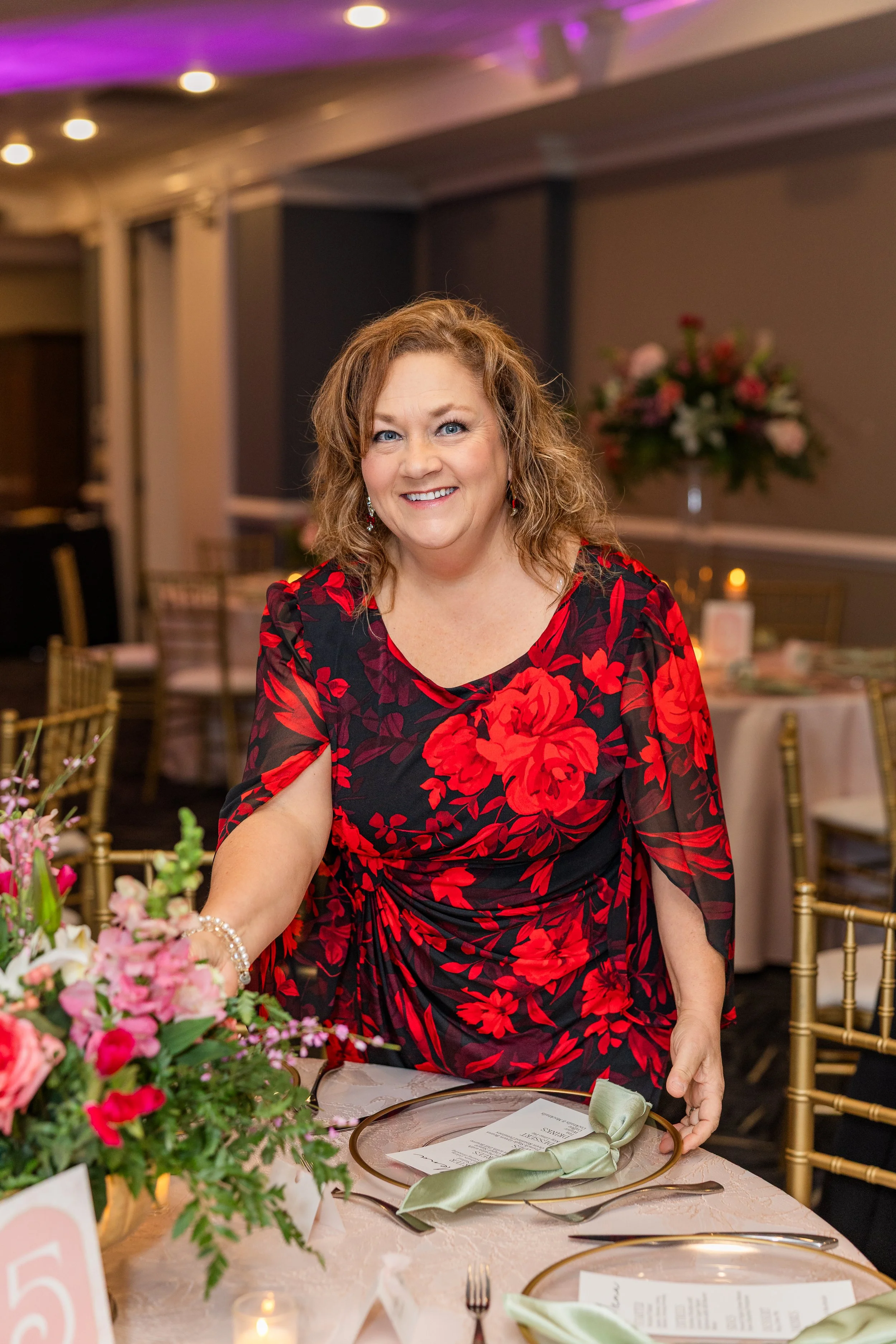A joyful woman with curly brown hair in a floral dress decorating a dinner table with flowers and table settings, in an elegant event space.
