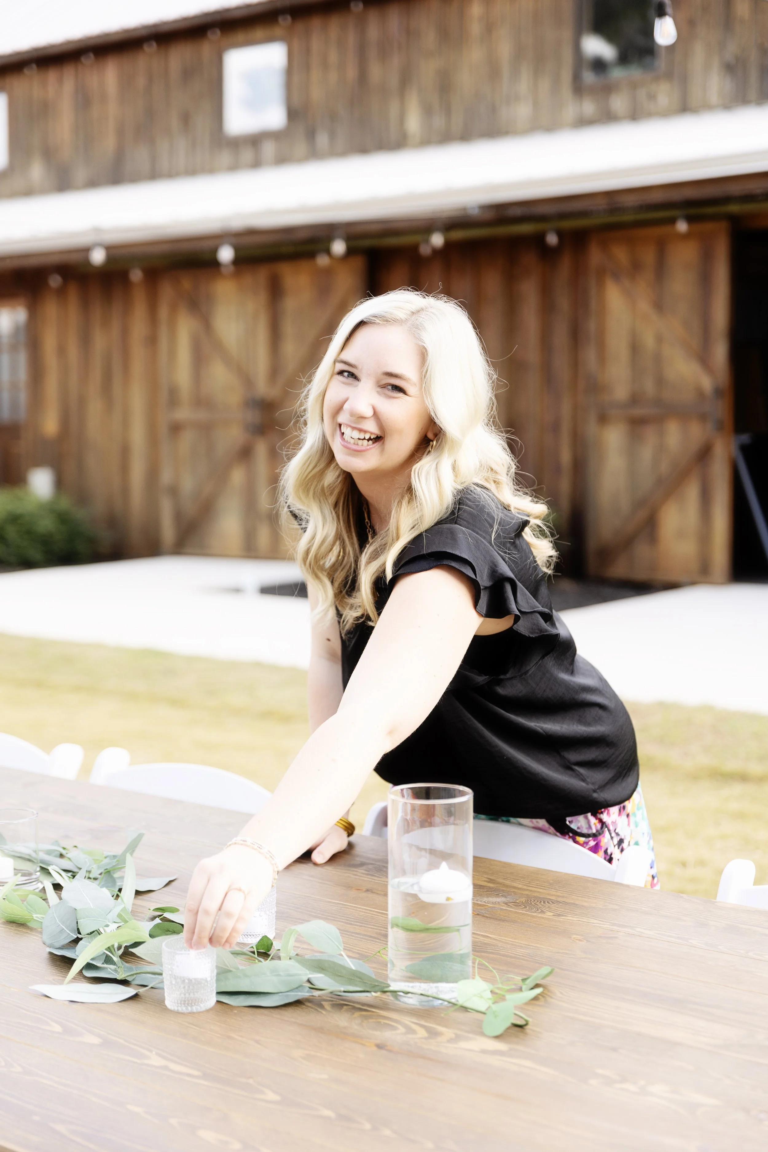 A smiling woman with blonde hair in a black top arranging greenery on a wooden table outdoors, with a rustic wooden barn in the background.