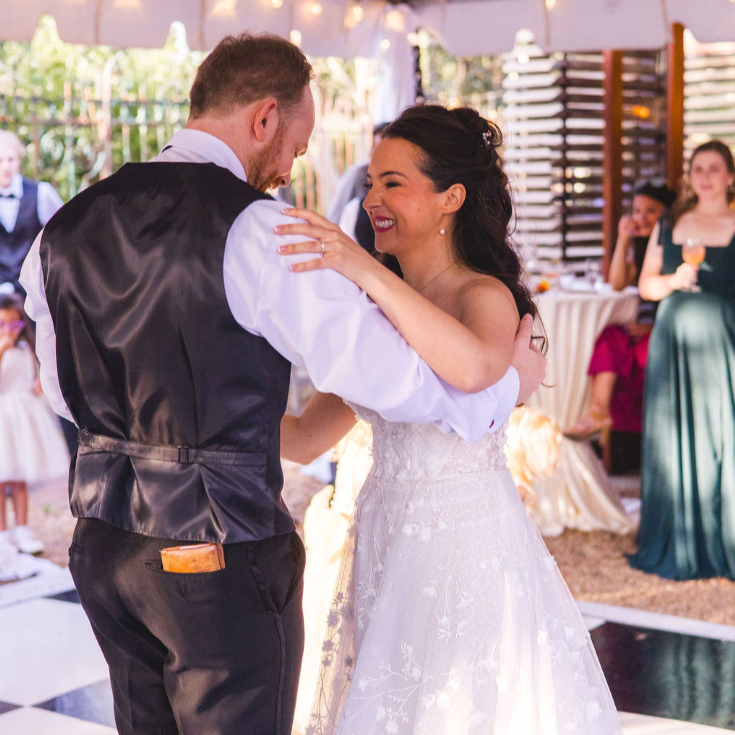 A bride and groom share a dance at their wedding reception, smiling and looking at each other, with guests in the background.