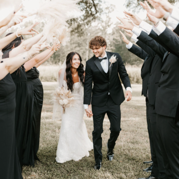 Bride and groom walking hand-in-hand through a tunnel of friends and family dressed in black, outdoors.