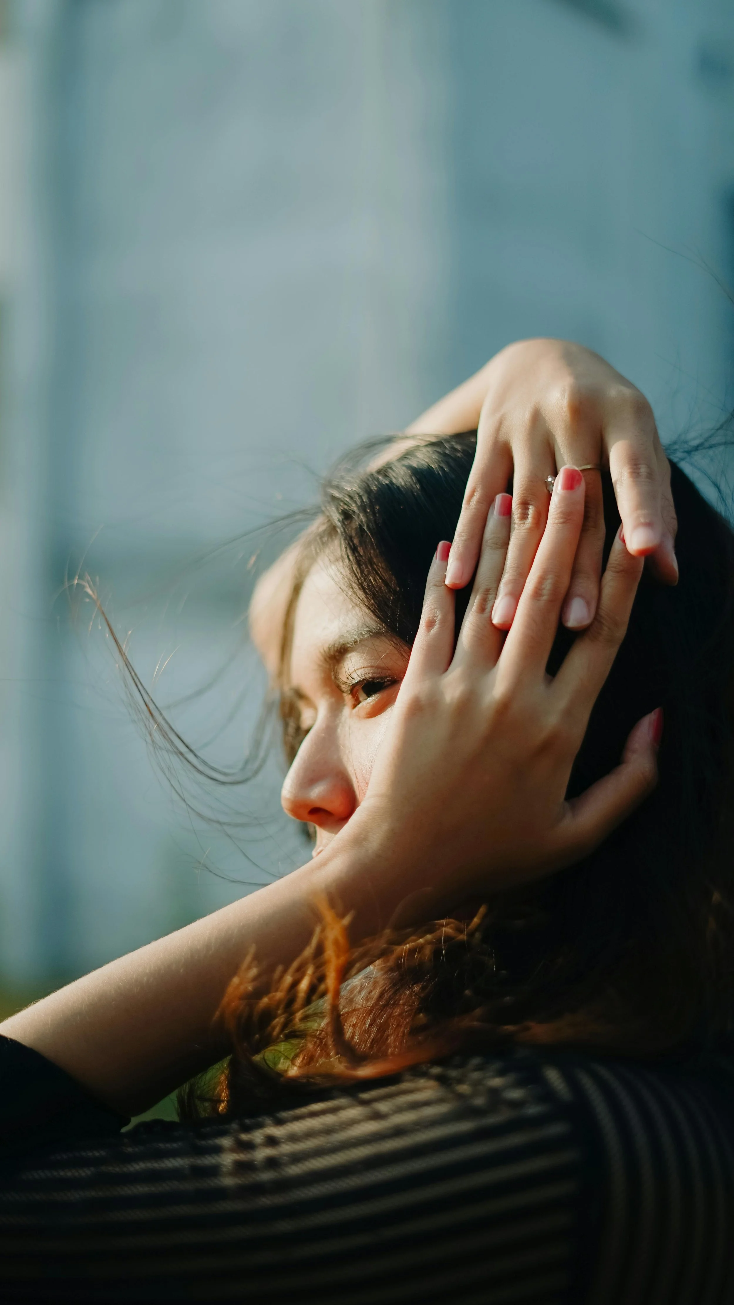 Close-up of a woman with dark hair, gently touching her head with one hand, eyes closed, sunlight illuminating her face, background blurred.