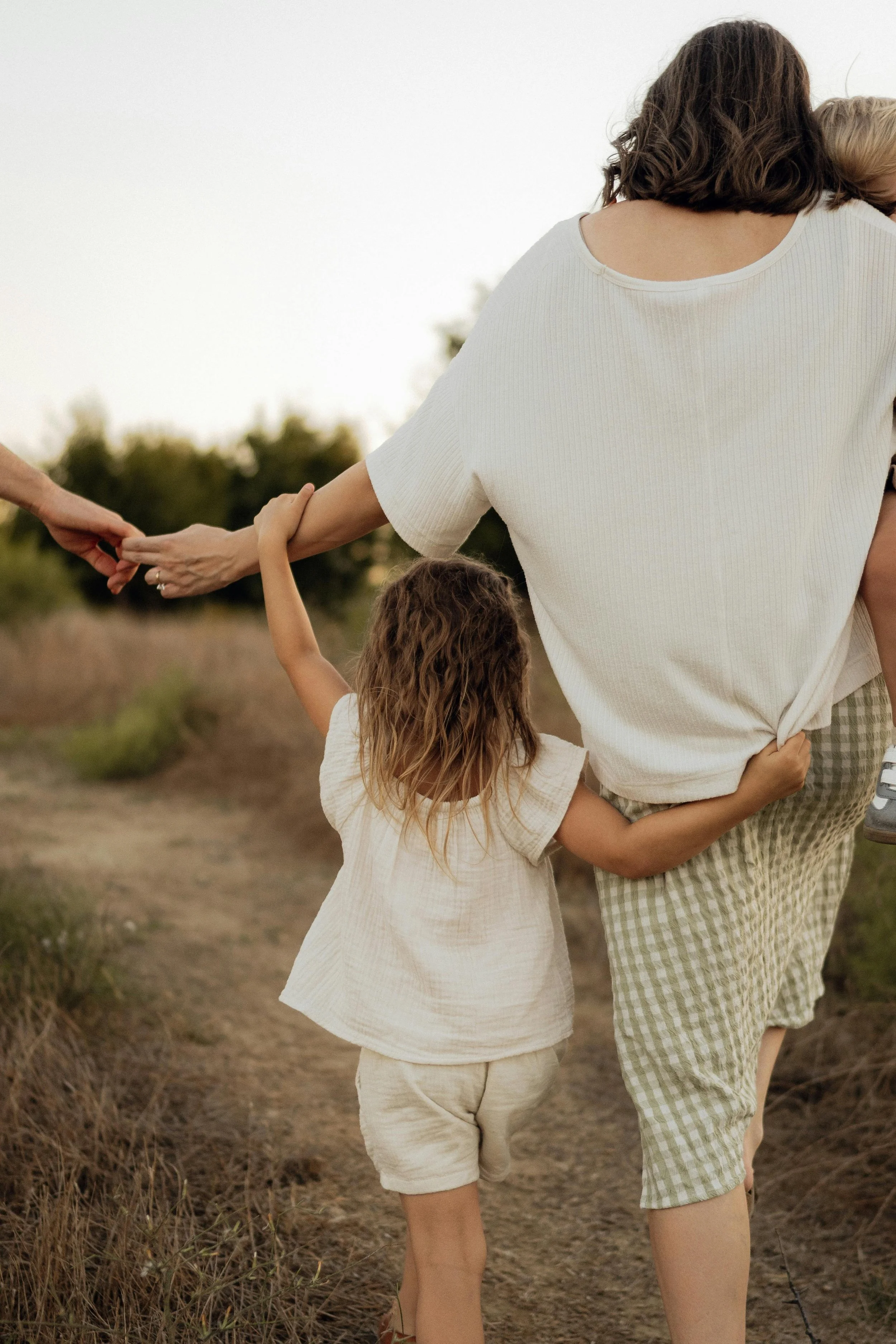 A woman holding hands with a young girl while walking outdoors on a dirt path, with trees in the background during sunset.