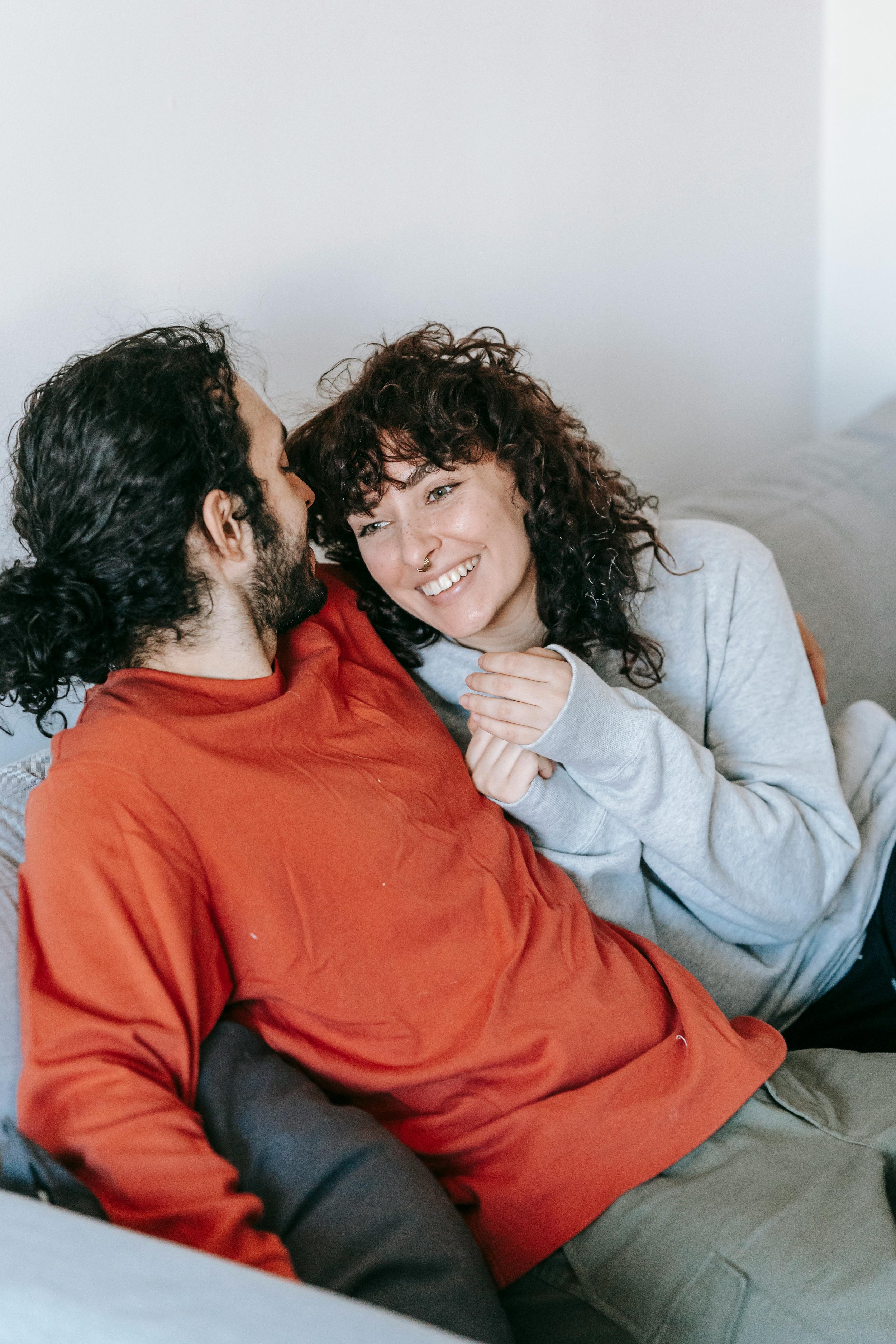 A man and woman sitting close together, smiling and looking at each other, in a cozy indoor setting.