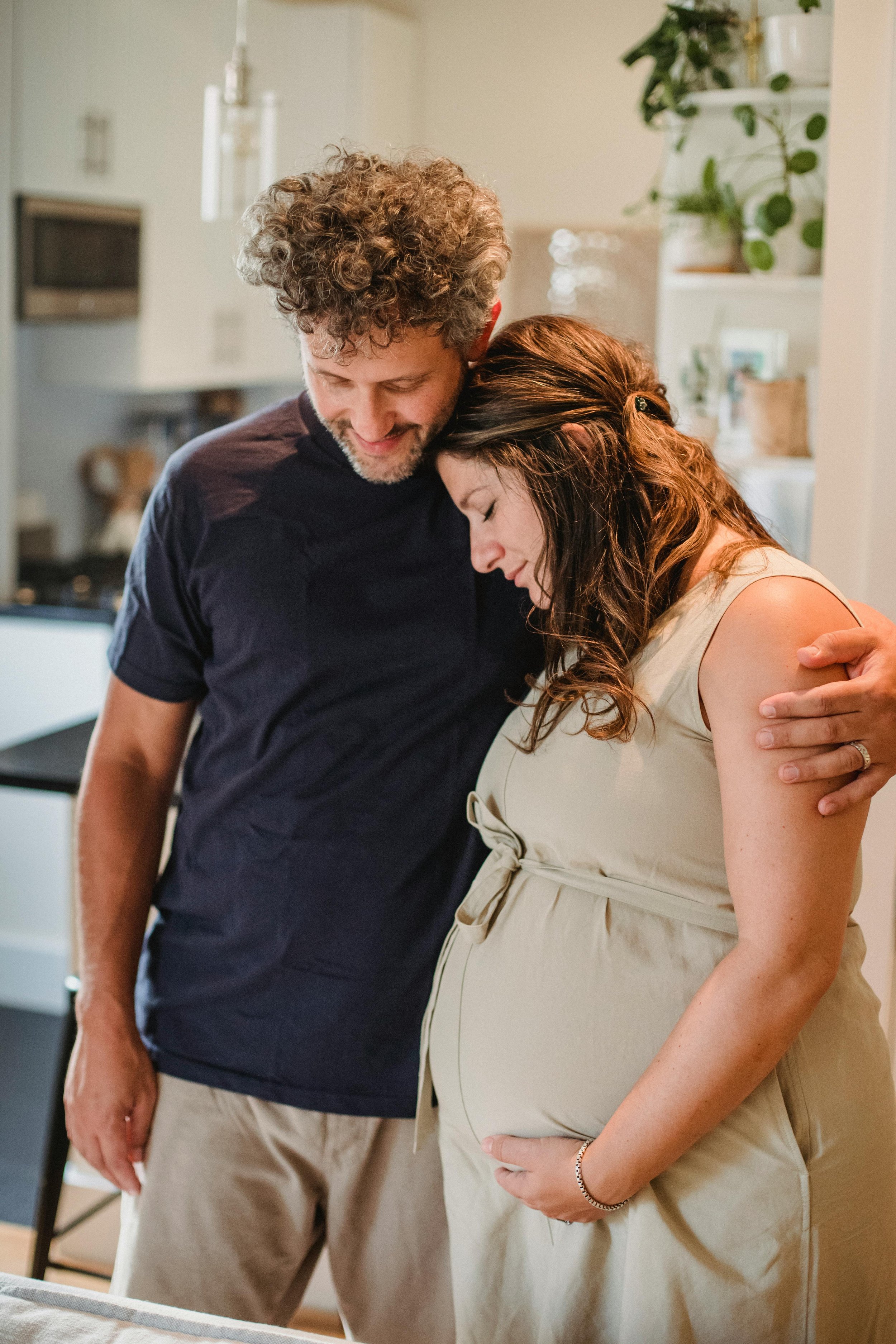 A man and pregnant woman share a tender moment, with the man gently resting his head on the woman's forehead and both smiling softly, in a cozy indoor setting.