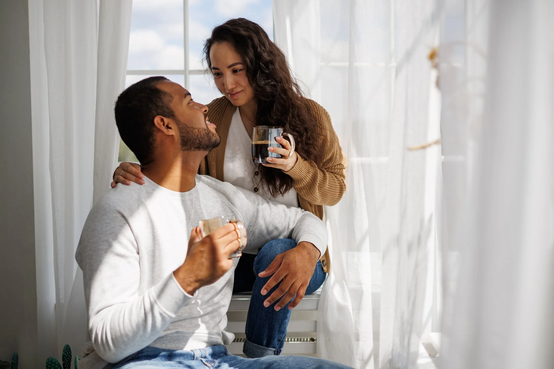 A woman with long dark hair and a man with short dark hair sitting by a window with white curtains. The woman is holding a glass cup of coffee or tea, and the man is holding a similar cup. They are smiling and looking at each other in a cozy, bright room.