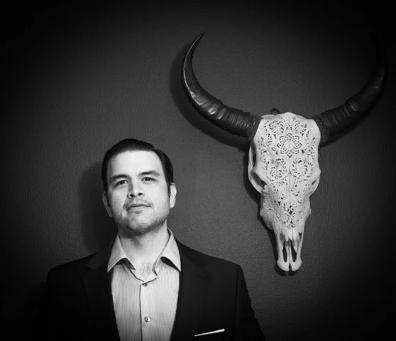 A man in a suit standing next to a decorated animal skull with large horns mounted on the wall.