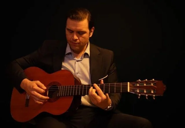 Man in a suit playing an acoustic guitar against a dark background.