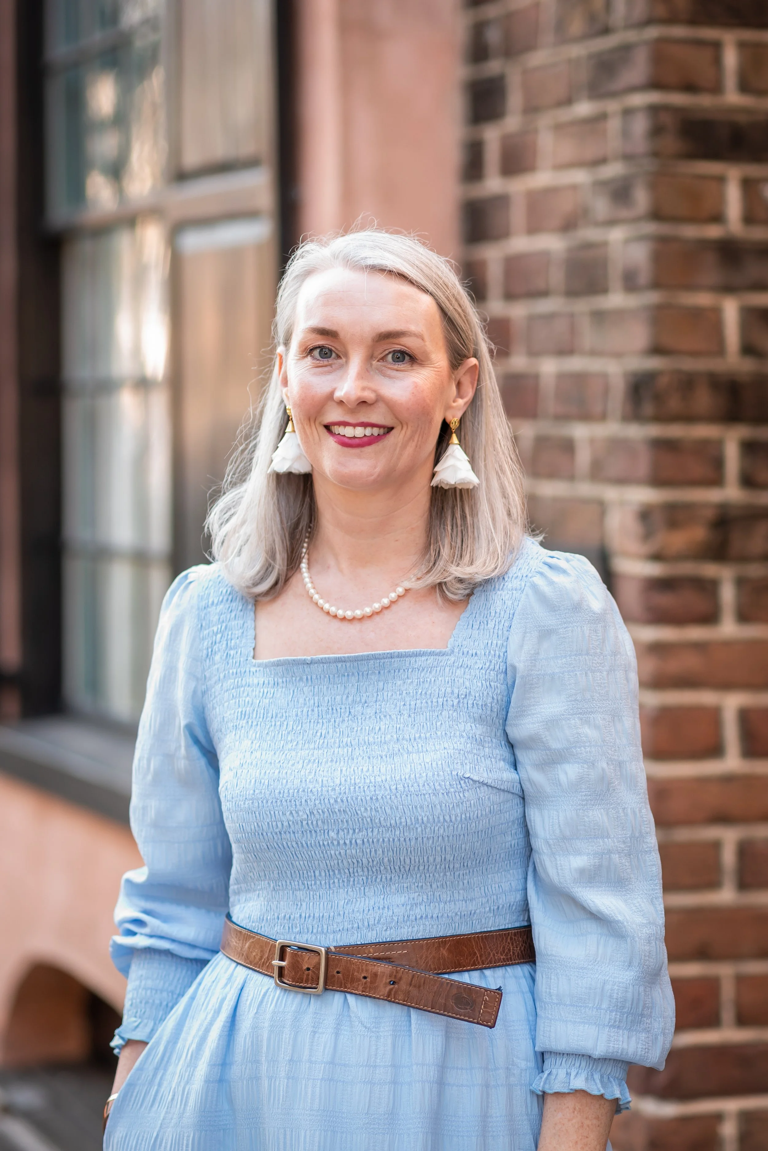 A woman with gray hair, wearing a light blue dress with a brown belt, pearl necklace, and white tassel earrings, standing outdoors in front of a brick building and window, smiling at the camera.