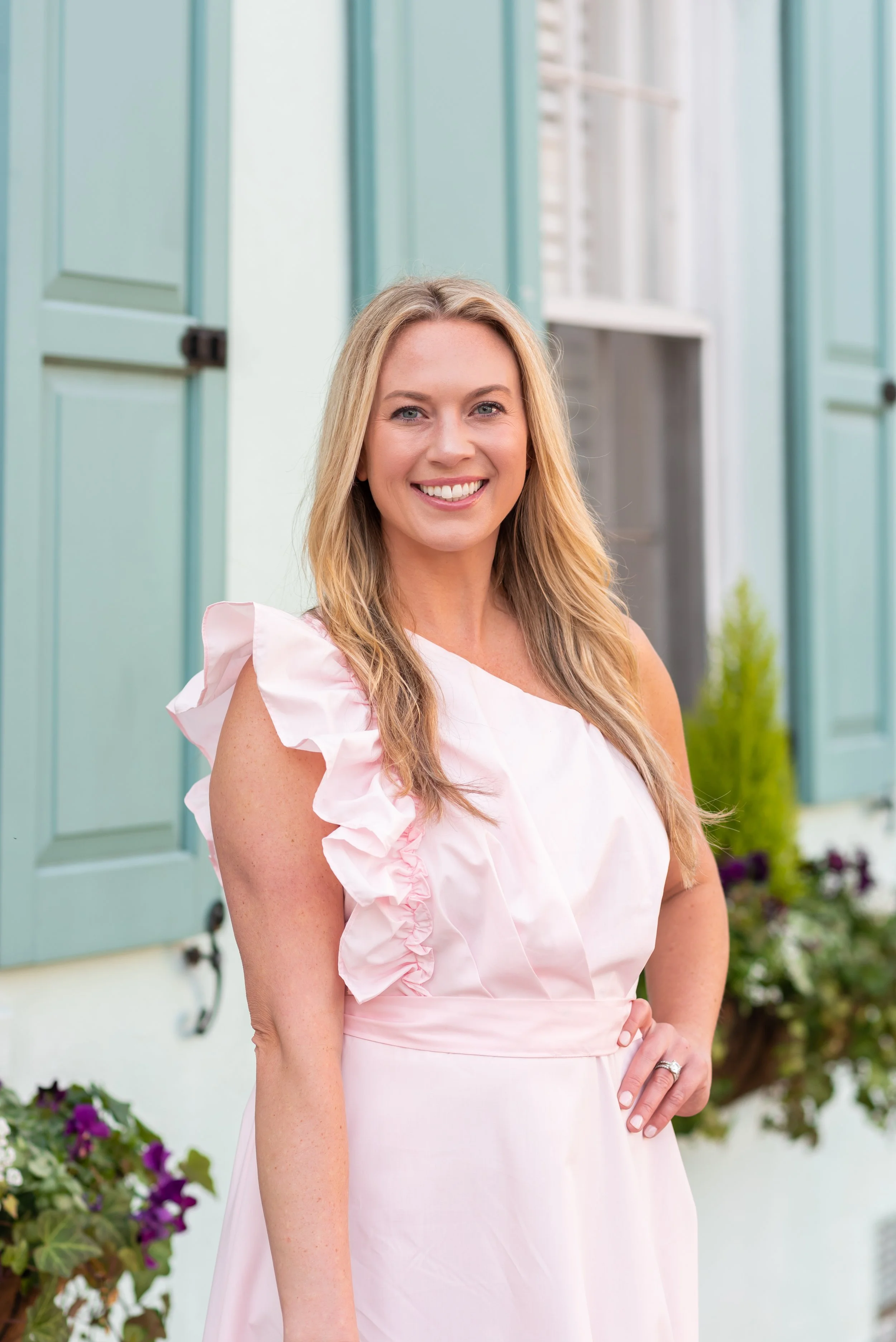 Smiling woman with blonde hair wearing a light pink dress standing outside near light teal window shutters with potted flowers nearby.