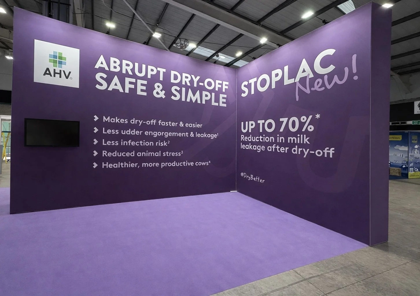 Trade show booth with purple walls promoting AHV's STOPLAC dry-off product, highlighting benefits like faster drying, less udder engorgement, lower infection risk, animal stress reduction, and healthier cows, with up to 70% reduction in milk leakage 