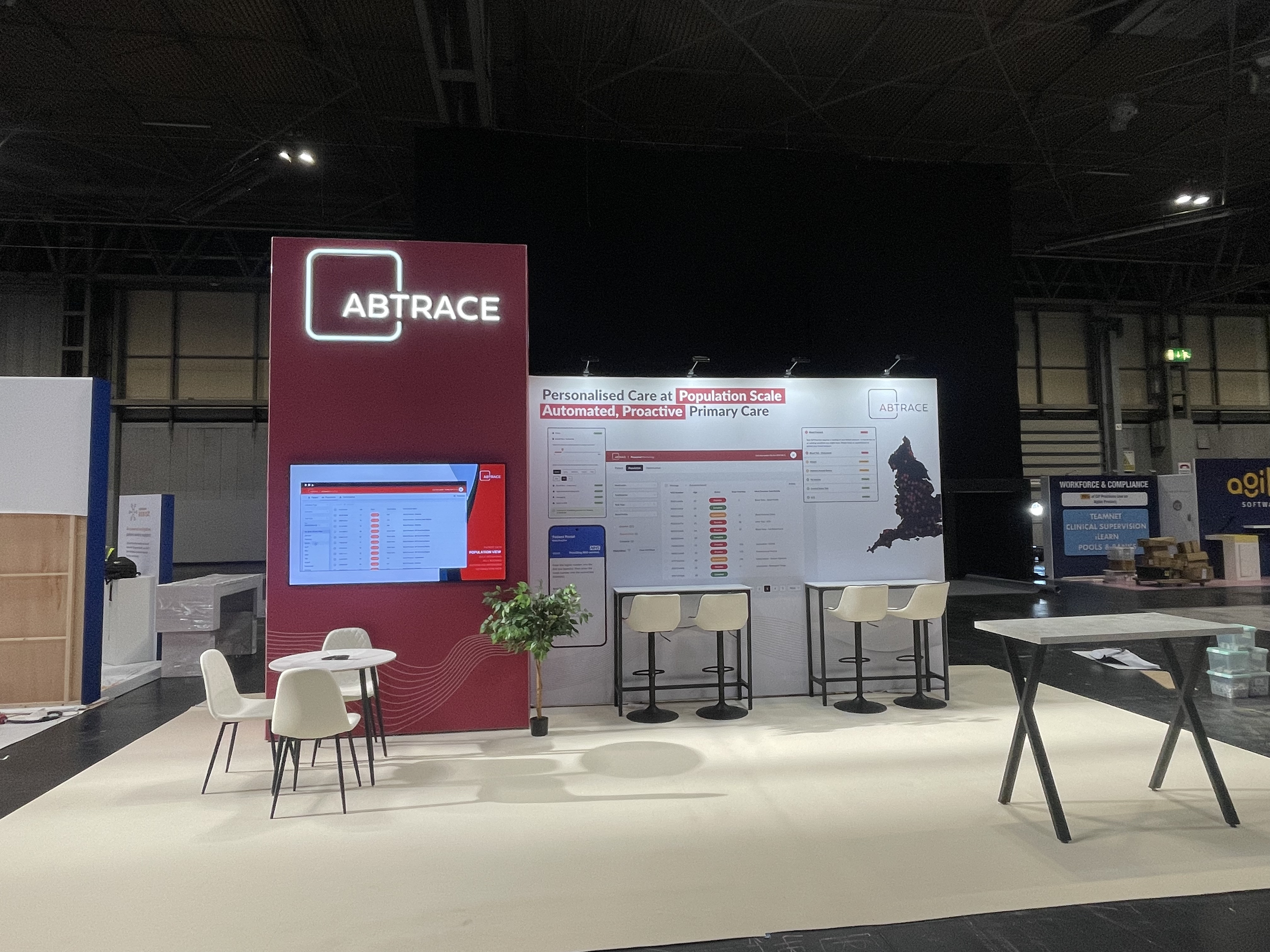 Trade show booth for ABTRACE with a red and white color scheme, featuring a large screen, a potted plant, a small round table with chairs, and a large poster displaying healthcare data and a map of England.