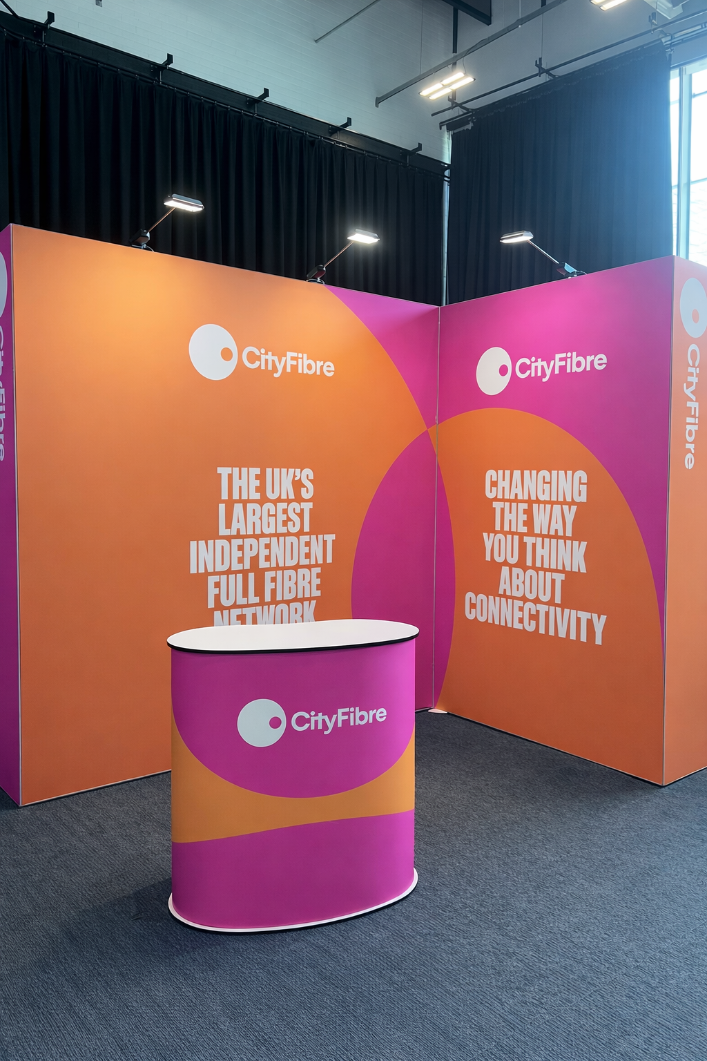 Colorful CityFibre exhibition booth with orange and pink backdrop, featuring the CityFibre logo, and a matching pink counter with the logo, promoting the UK's largest independent full fibre network and connectivity ideas.