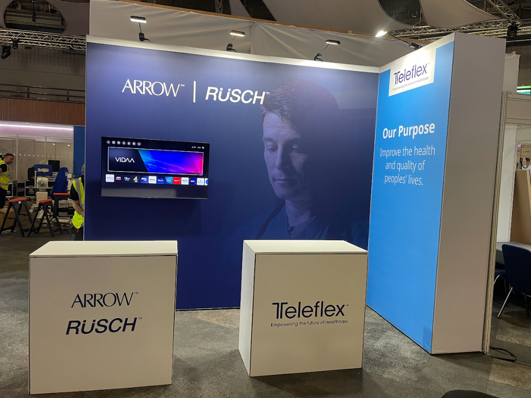 Trade show booth for Arrow Rusch and Teleflex featuring a large display screen, a backdrop with a medical professional's face, and the company's purpose to improve health and quality of life.