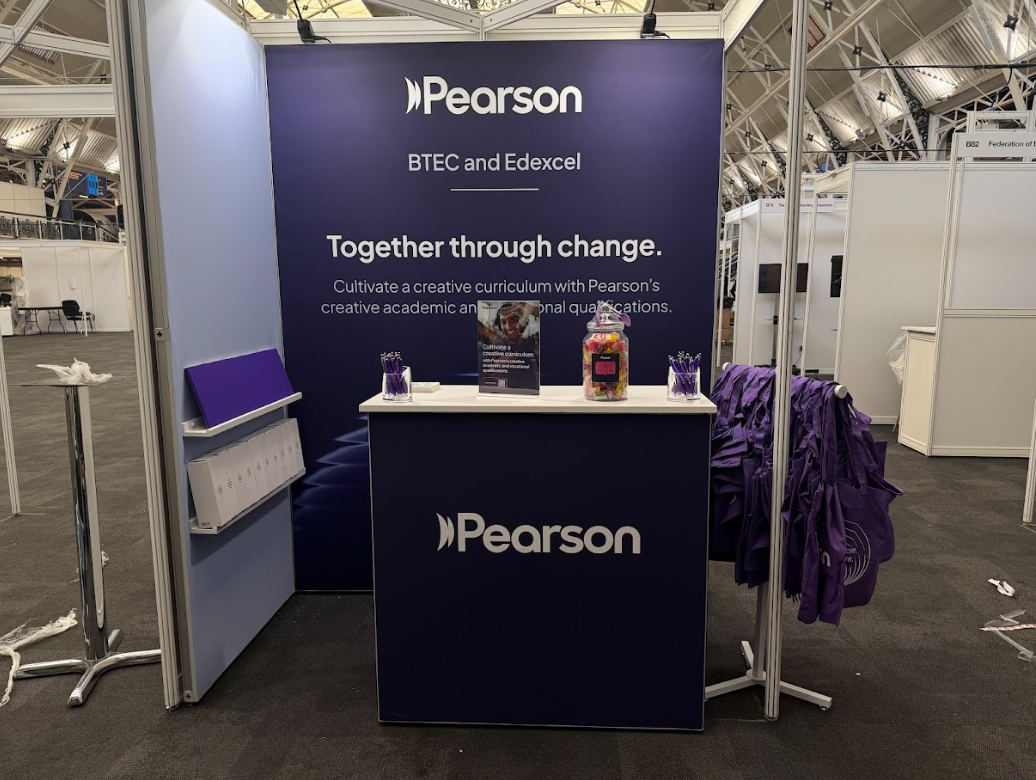 Pearson booth at an exhibition, featuring a large purple backdrop with the Pearson logo and the text 'BTEC and Edexcel. Together through change.' A white counter with purple pens, a candy jar, and promotional materials. Purple shirts hanging on a sta