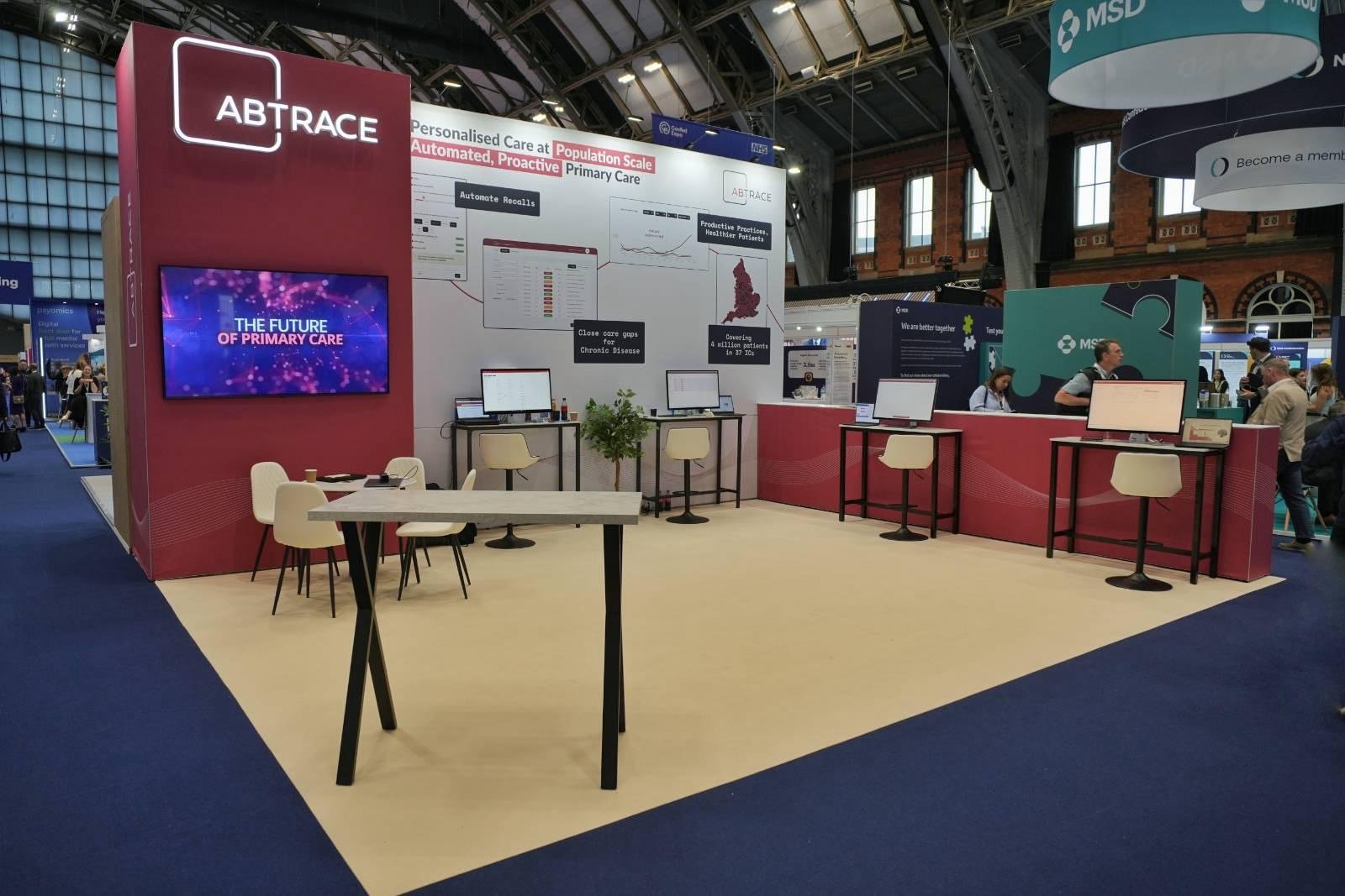 Trade show booth for ABTRACE featuring monitors, chairs, and informational signage about primary care technology.