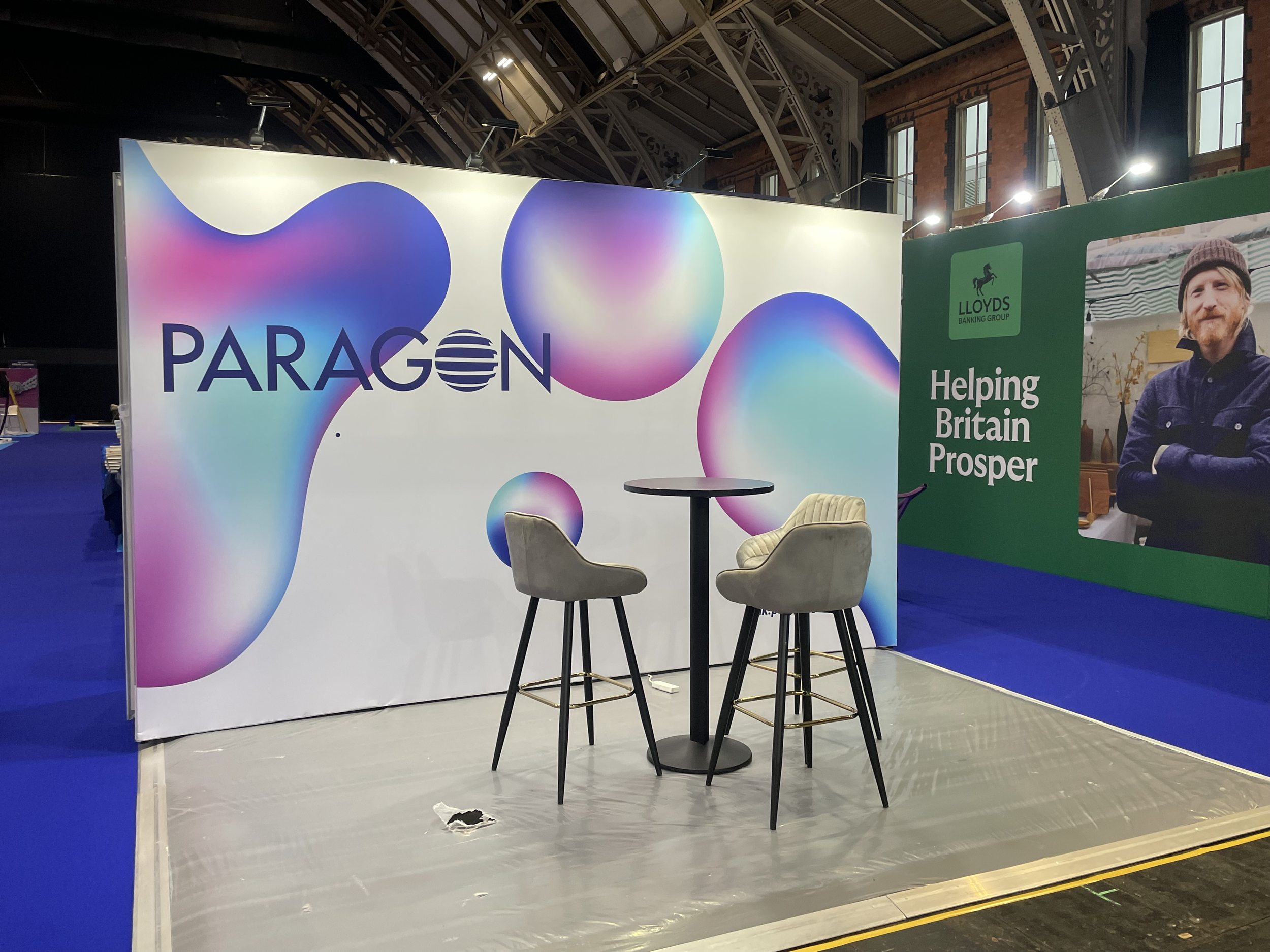An exhibition booth with the word "PARAGON" on a colorful abstract background, featuring two high chairs and a small round table in front of it, with a large poster on the side that reads "Helping Britain Prosper" and a picture of a man.