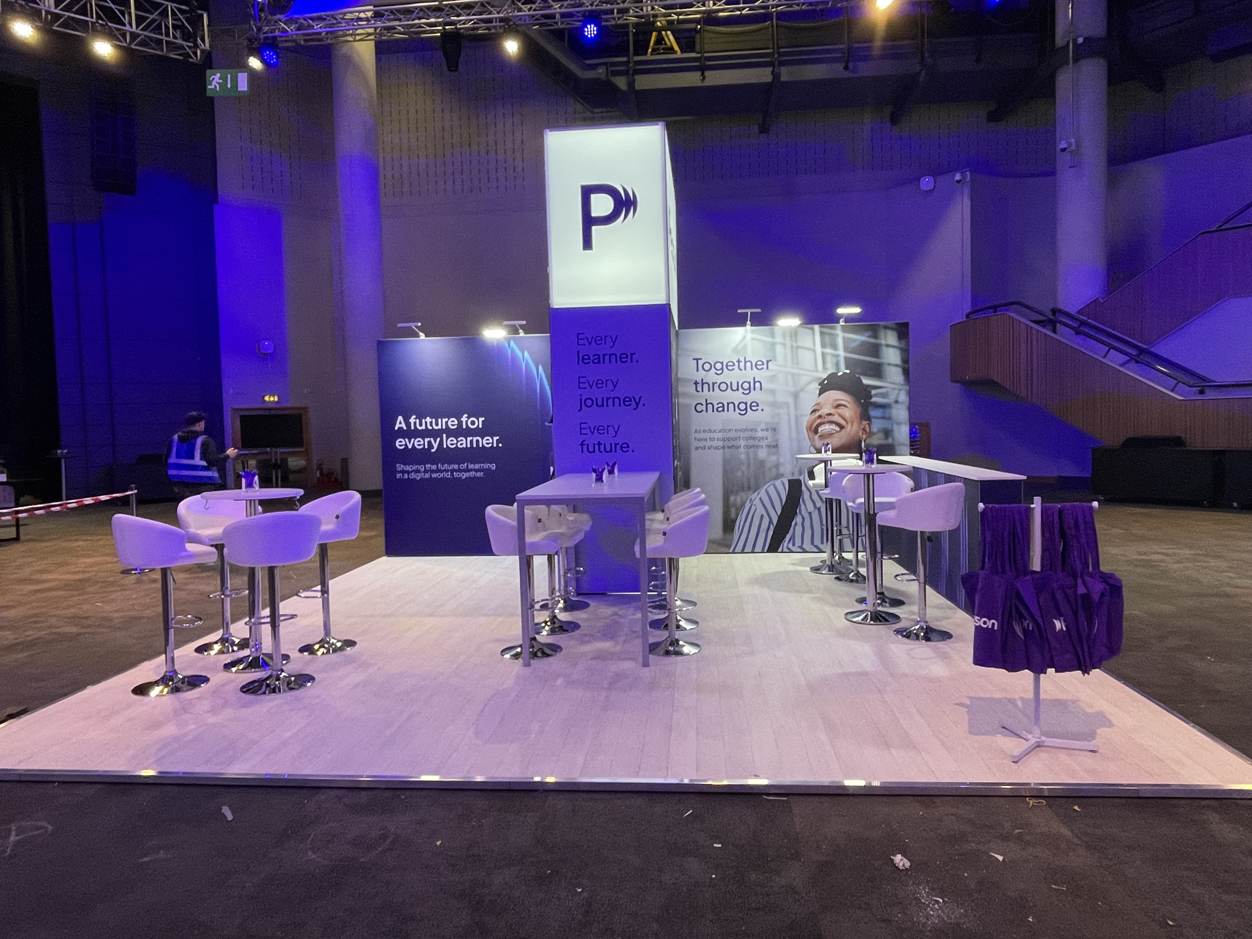 Event booth with purple and blue branding, featuring tables and A-frame displays, in a large indoor space with high ceilings and staircase.