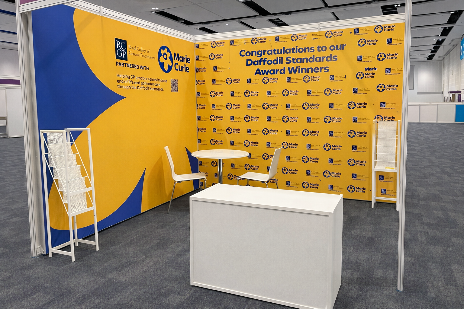 Exhibition booth with yellow and blue backdrop featuring Marie Curie branding and congratulatory message for Daffodil Standards Award winners, with two white chairs, a small round white table, and a white display rack.