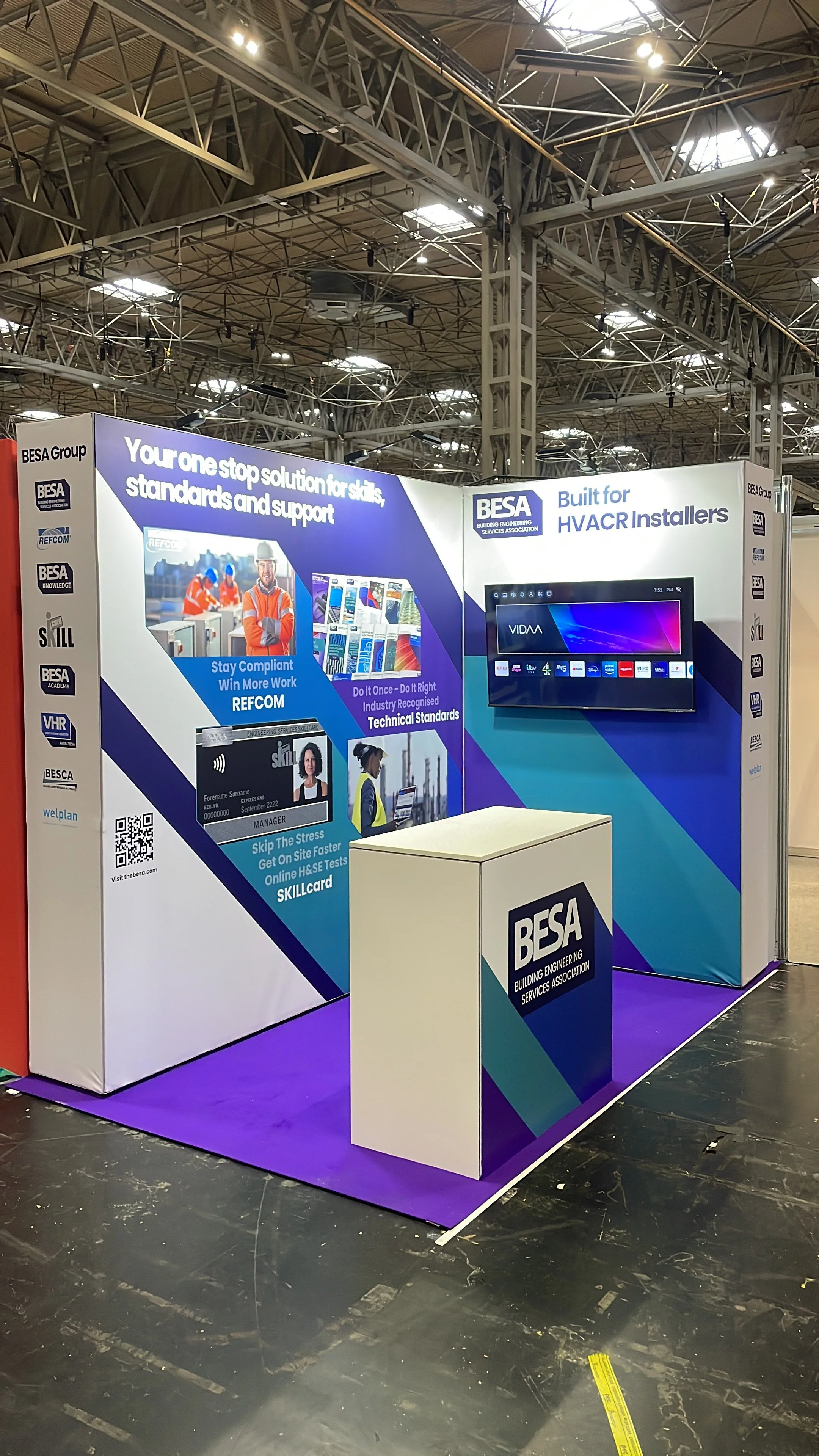 Trade show booth for BESA (Building Engineering Services Association) with displays of industry standards, software, and technical information. Features a monitor, informational posters, and a podium, set on a purple carpet in a large exhibition hall
