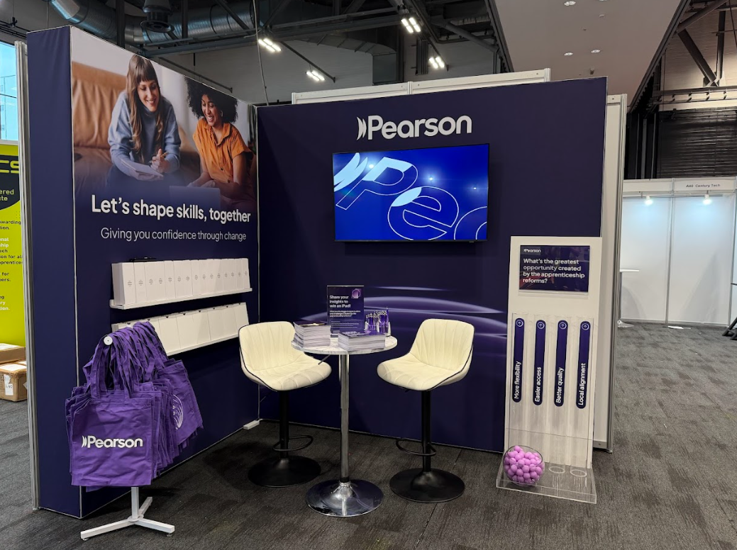 A Pearson booth at an expo with a dark blue backdrop. The booth features a large screen, promotional materials, and a table with two white chairs. There are purple tote bags hanging on a stand and a clear container with purple balls. The booth promot