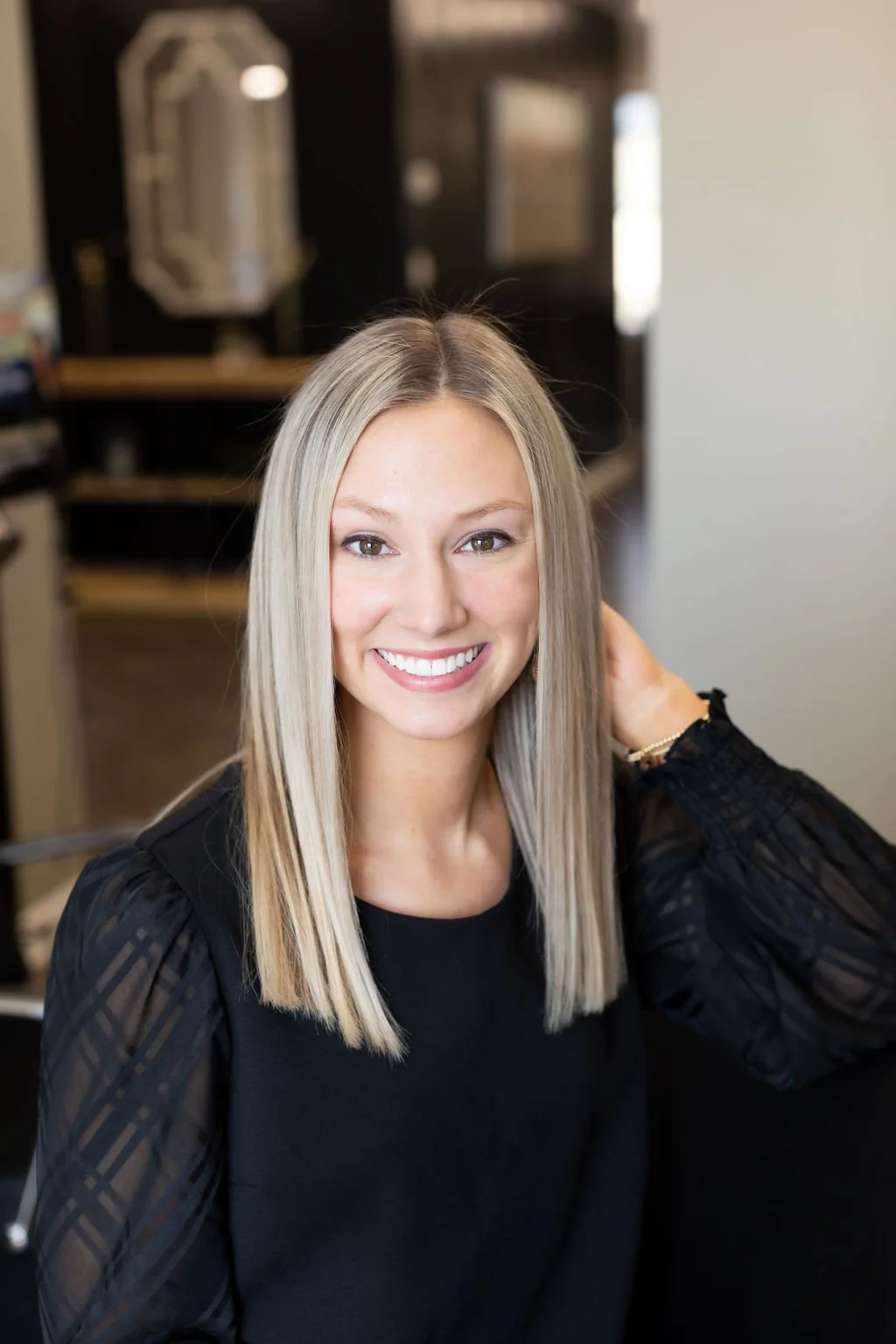 A woman with straight blonde hair smiling at the camera. She is wearing a black top with sheer, striped sleeves and is in an indoor setting with a blurred background.