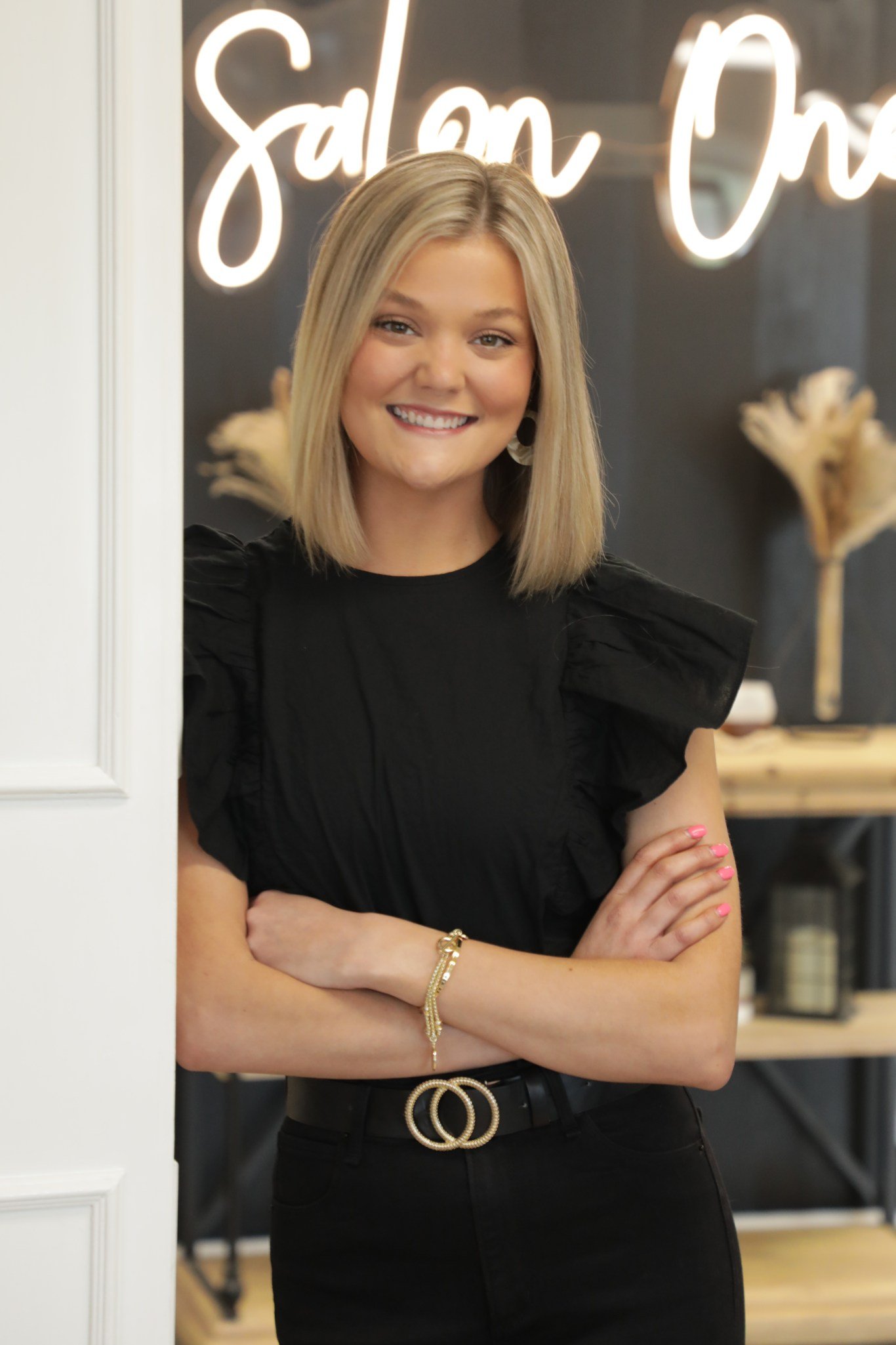 A woman with blonde hair smiling, standing with arms crossed, wearing a black dress and jewelry, in a modern interior with a neon sign and decorative plants.