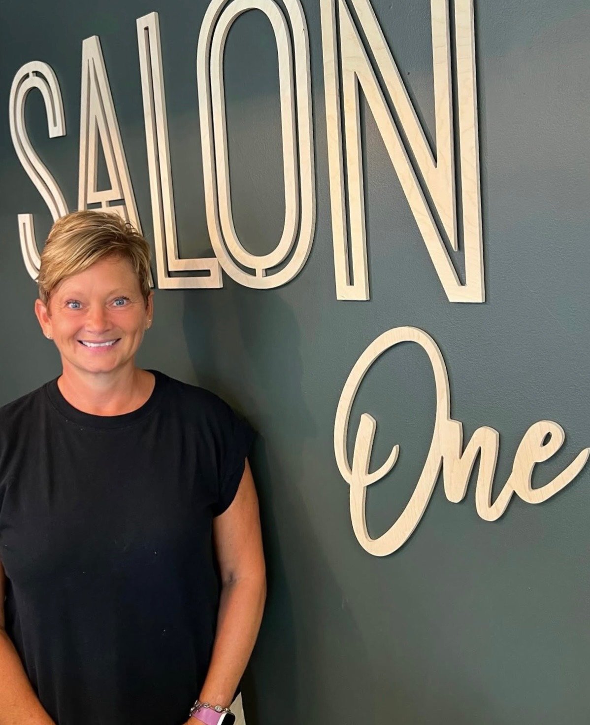 A smiling woman with short blond hair stands in front of a wall with large sign letters spelling 'SALON'. The wall color is dark teal.