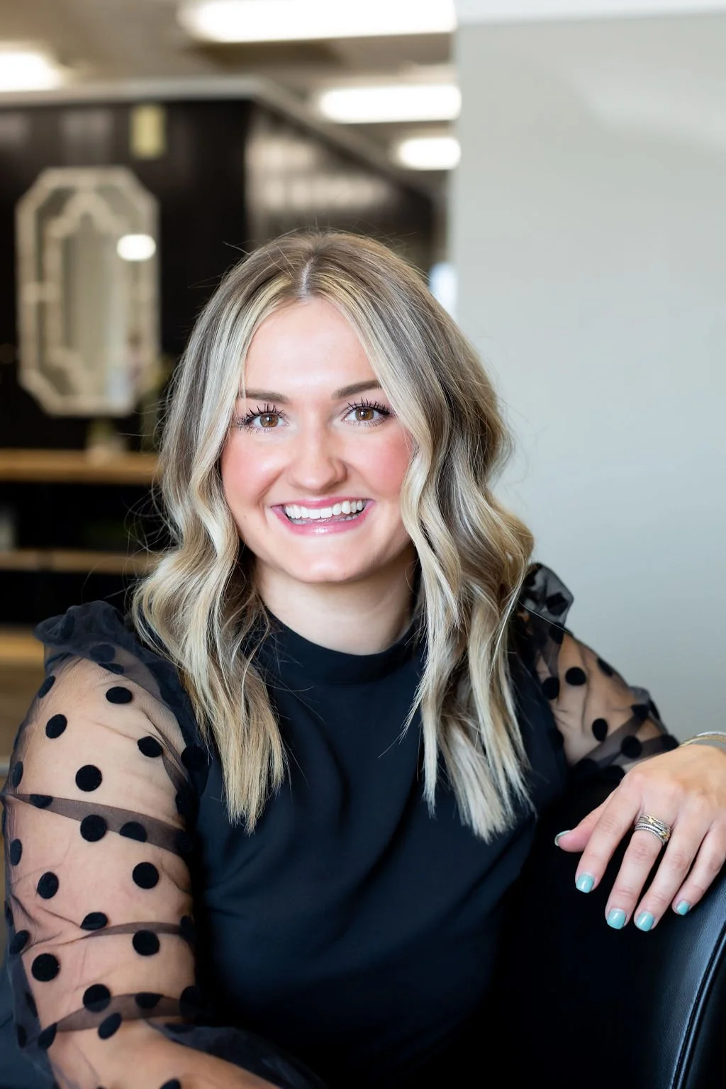 A smiling woman with shoulder-length blonde hair, wearing a black top with sheer polka-dot sleeves, sitting in an indoor setting.