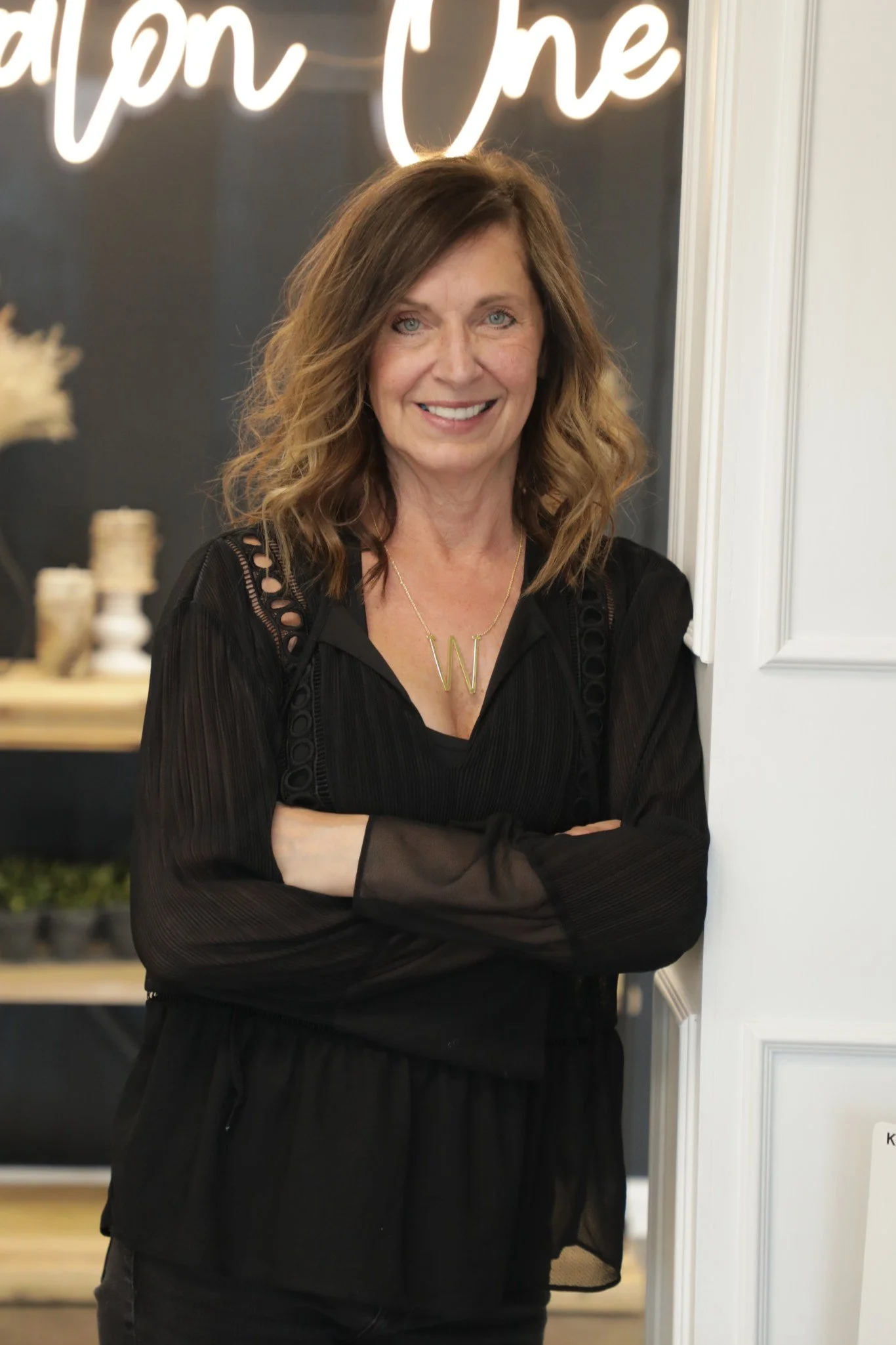 A woman with shoulder-length wavy brown hair, wearing a black blouse with lace details and a gold necklace with a 'W' pendant, smiling and standing with arms crossed inside a room with a dark wall and neon sign in the background.