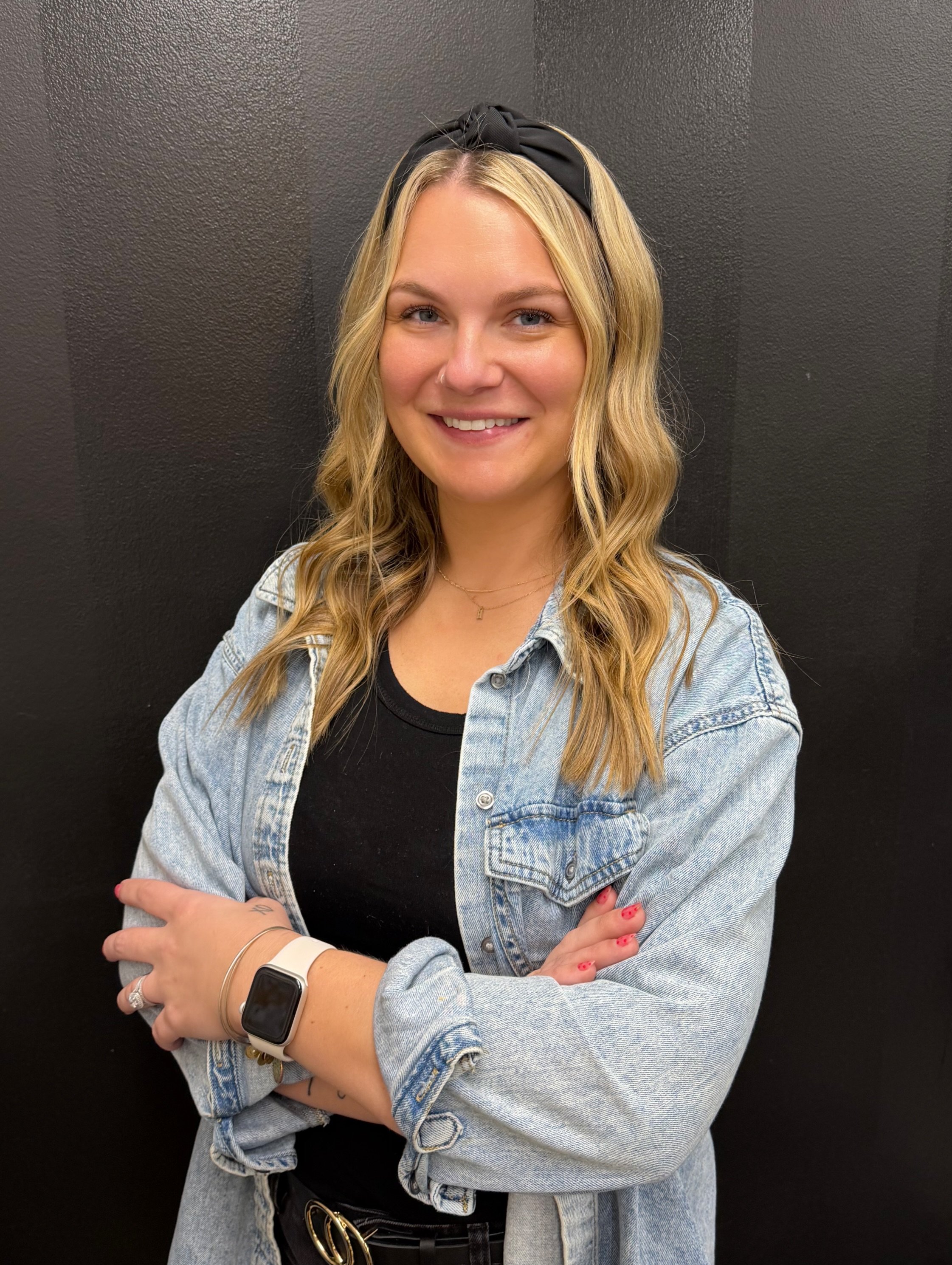 A young woman with blonde wavy hair wearing a black hairband, a black t-shirt, a light-wash denim jacket, and jewelry, smiling with arms crossed against a black background.