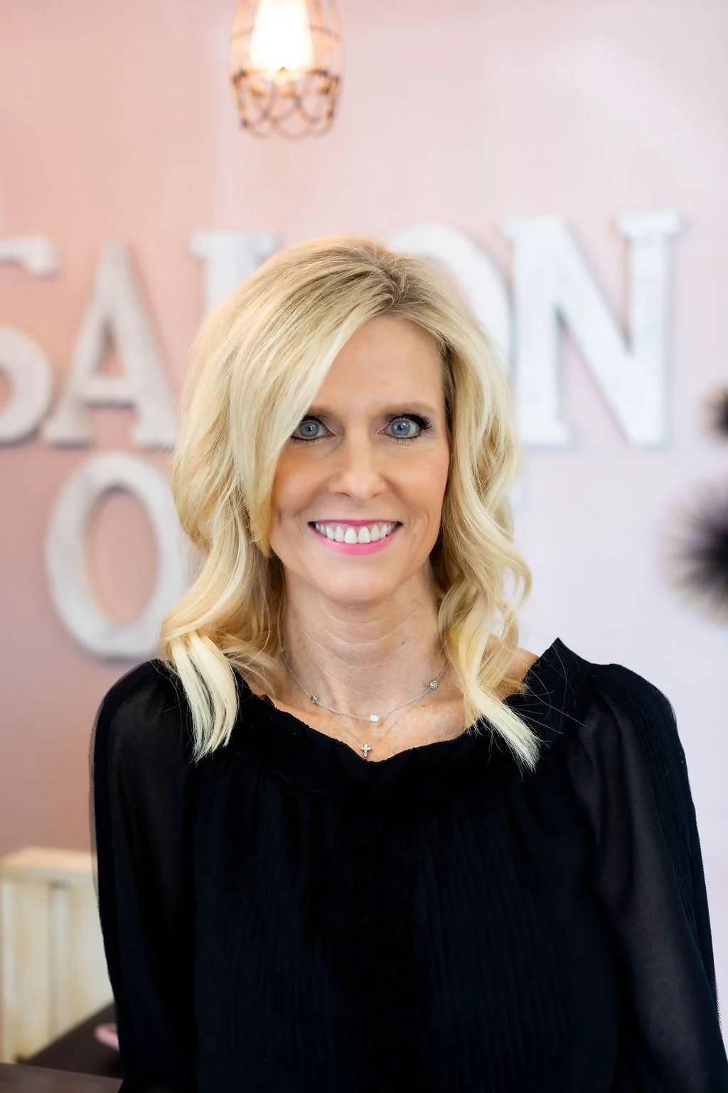 A woman with blonde hair and blue eyes smiling, wearing a black top, standing in front of a pink wall with large white letters and a hanging light fixture.