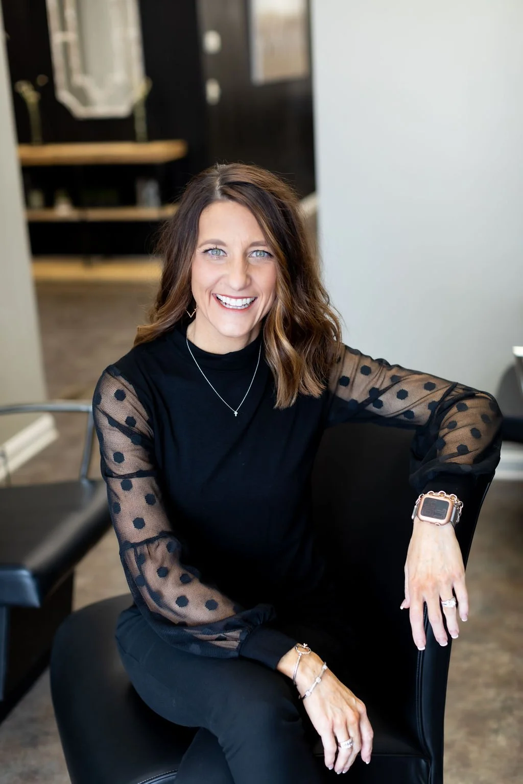 A woman with shoulder-length brown hair, blue eyes, and a bright smile sitting in a black chair. She is wearing a black top with sheer polka-dot sleeves, jewelry including a necklace, bracelets, and rings, and a smartwatch on her left wrist. She is indoors, with a blurry background that includes a mirror, a counter, and a black door.