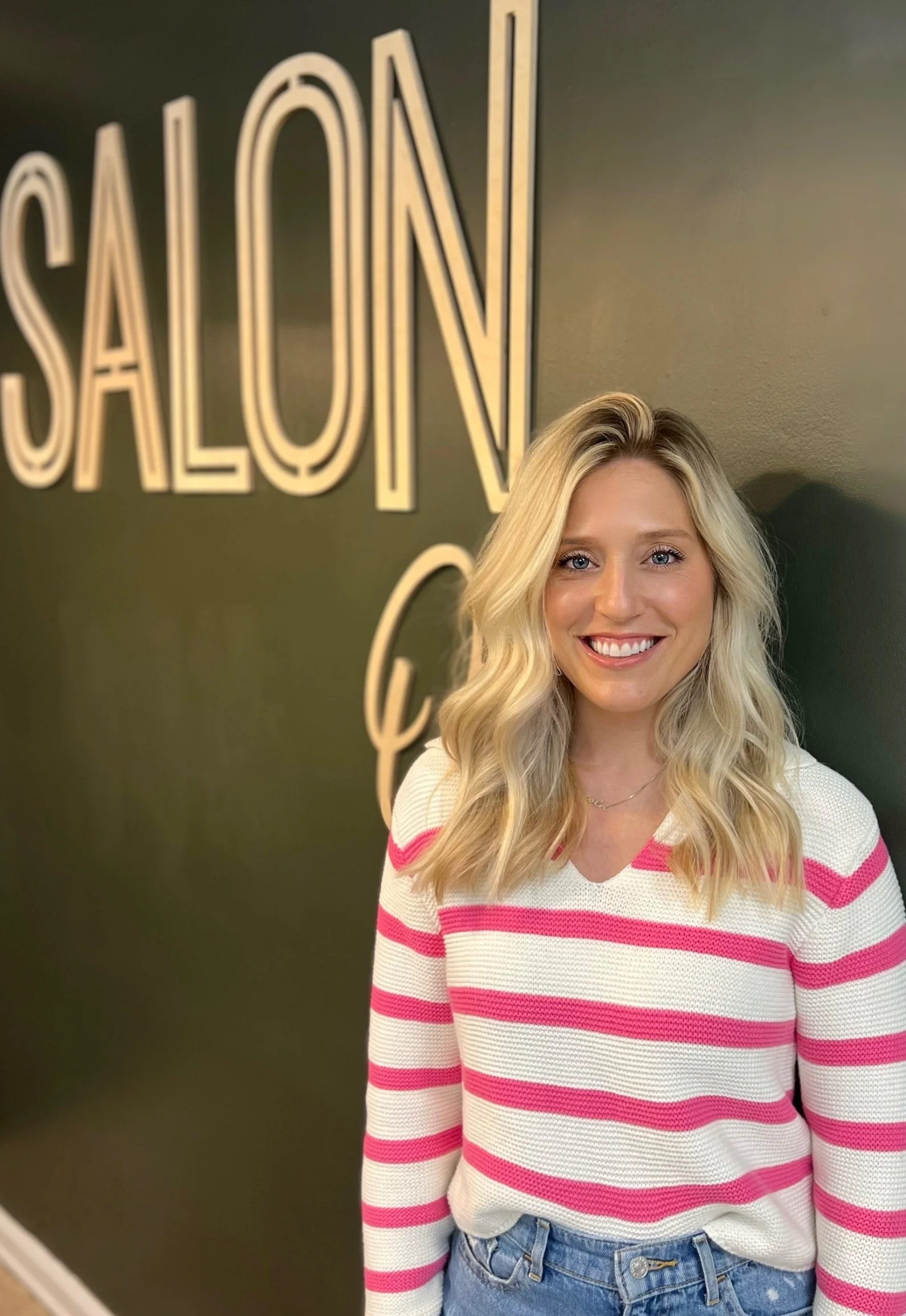 A young woman with blonde hair wearing a white and pink striped sweater and jeans, smiling and standing in front of a dark green wall with large illuminated word 'SALON'.