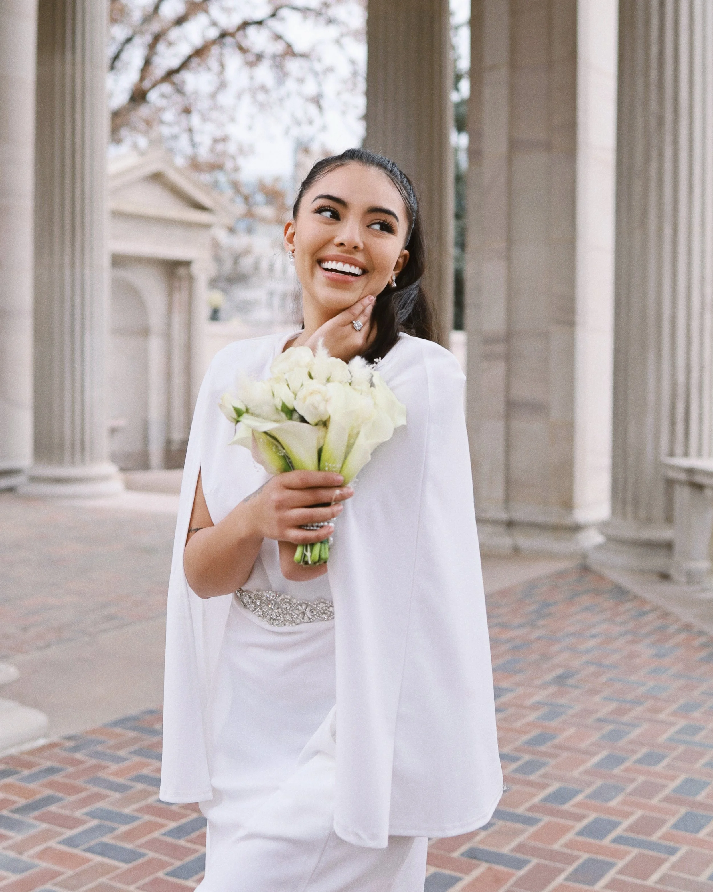 Young woman in white wedding dress holding a bouquet of white flowers, smiling, standing outside with columns and a brick pathway.