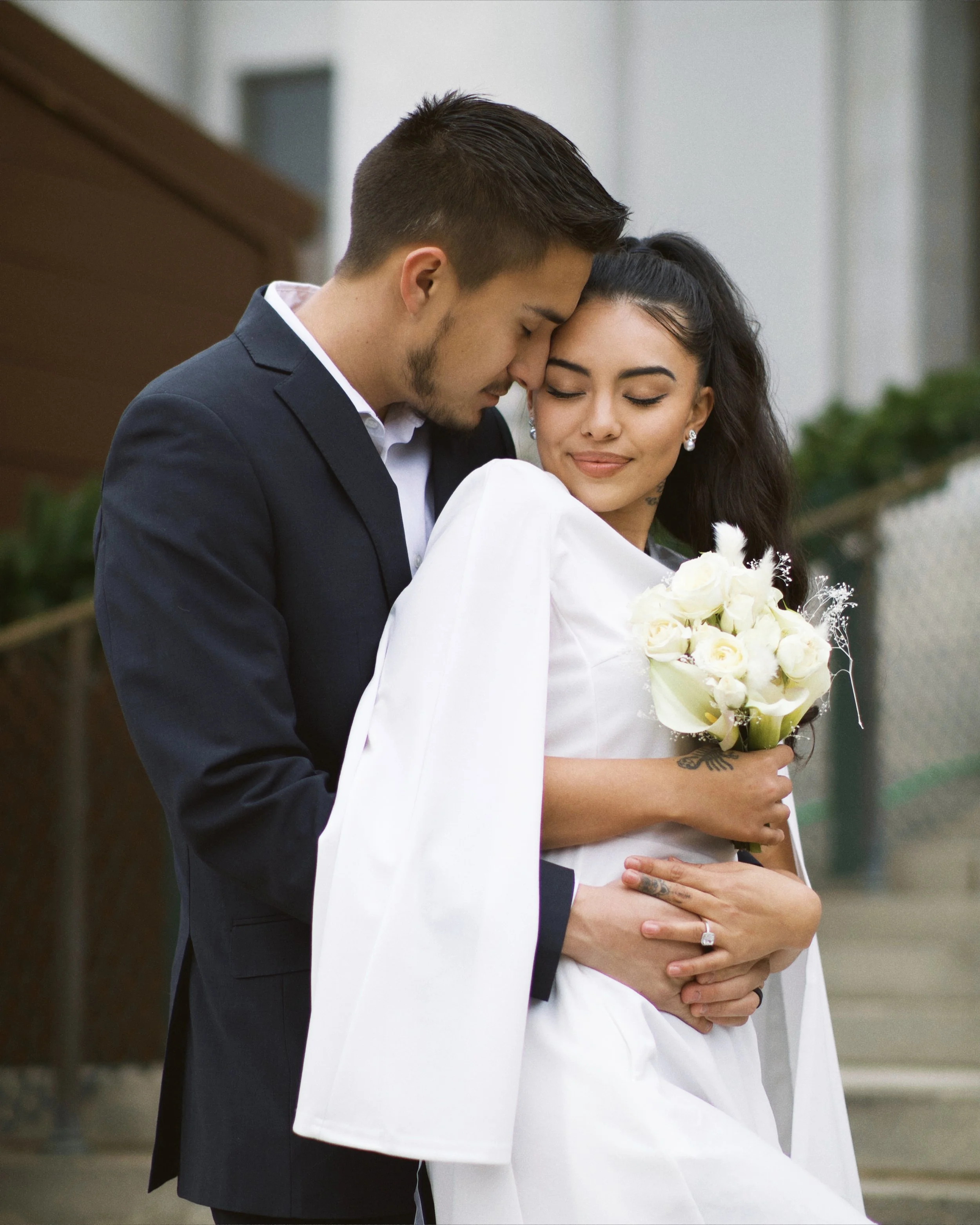 A couple dressed in wedding attire, the man in a dark suit and the woman in a white wedding dress, holding a bouquet of white roses, embracing closely with their foreheads touching.