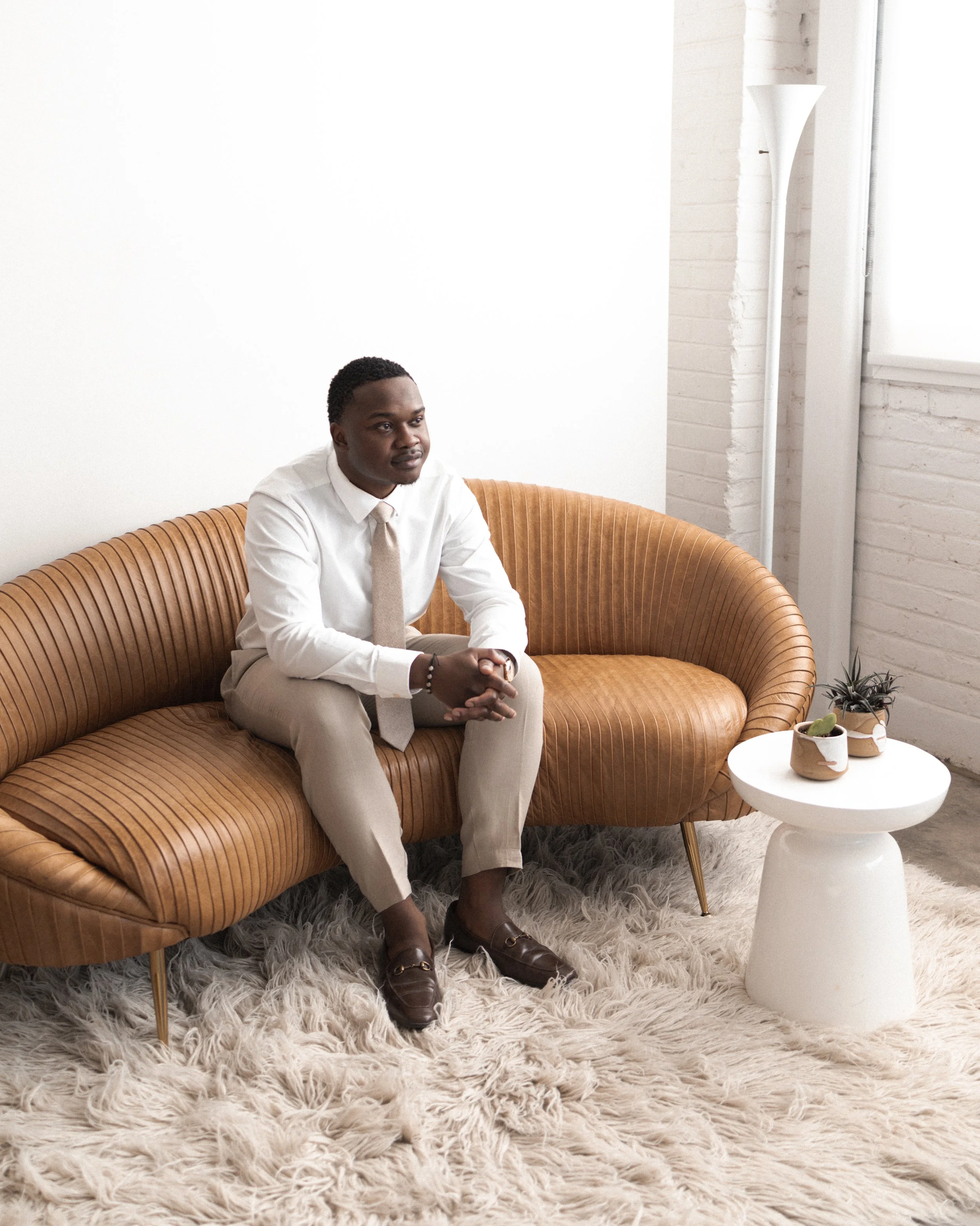 A man sitting on a tan, curved leather sofa in a bright, minimalist room with white walls and a shaggy off-white rug. There is a white side table with potted plants next to him.