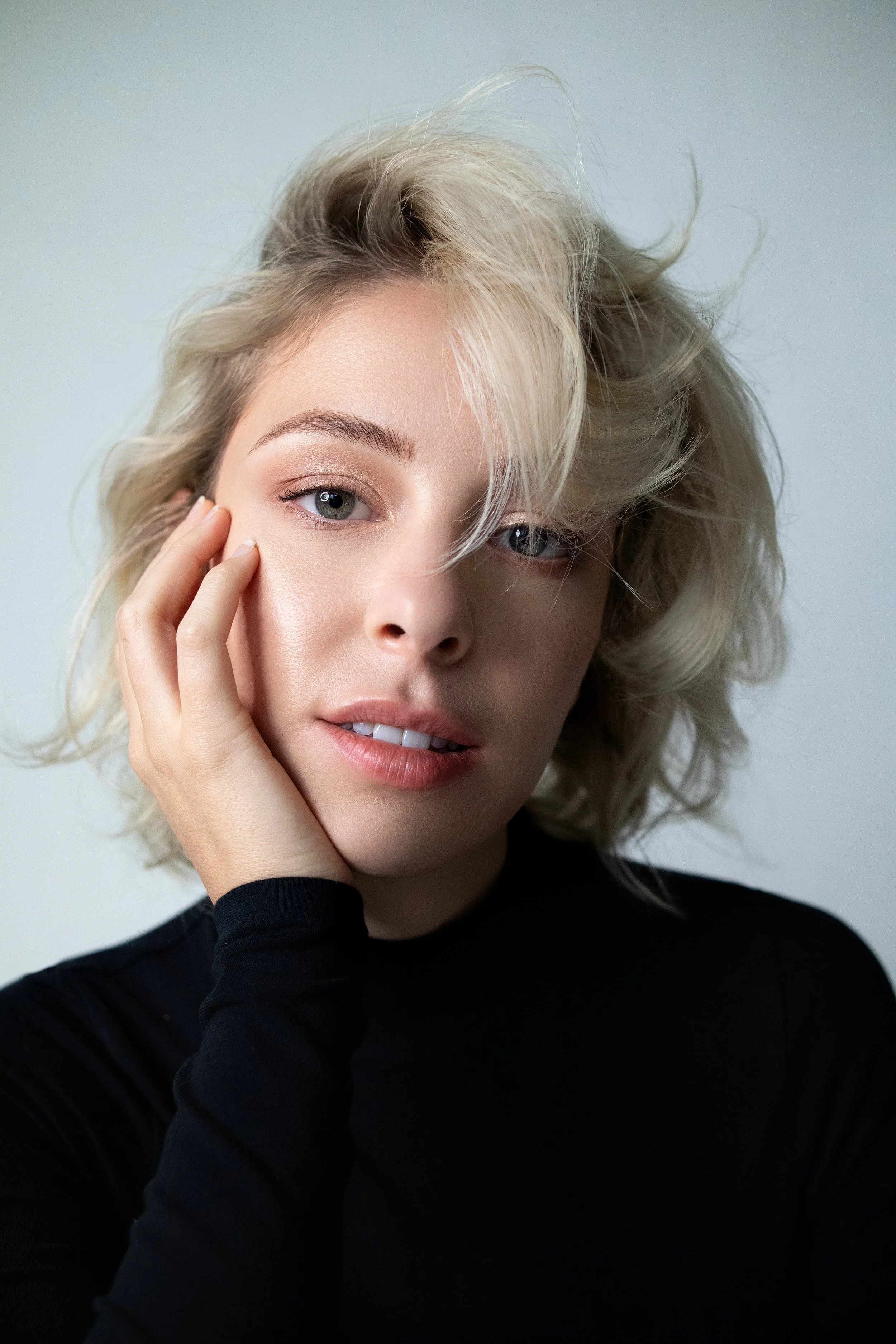 A woman with blonde, curly hair and blue eyes is touching her face with her left hand, wearing a black top, against a light grey background.