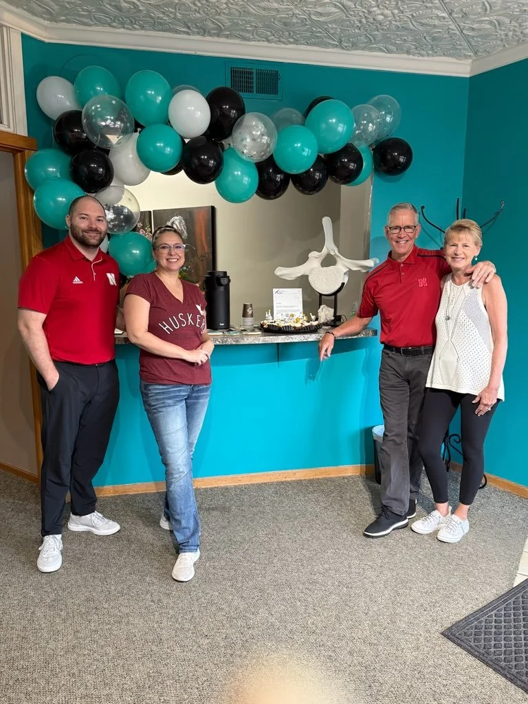 Four people standing in front of a balloon arch and a decorated table at a celebration or event in a room with teal walls and decorative ceiling.