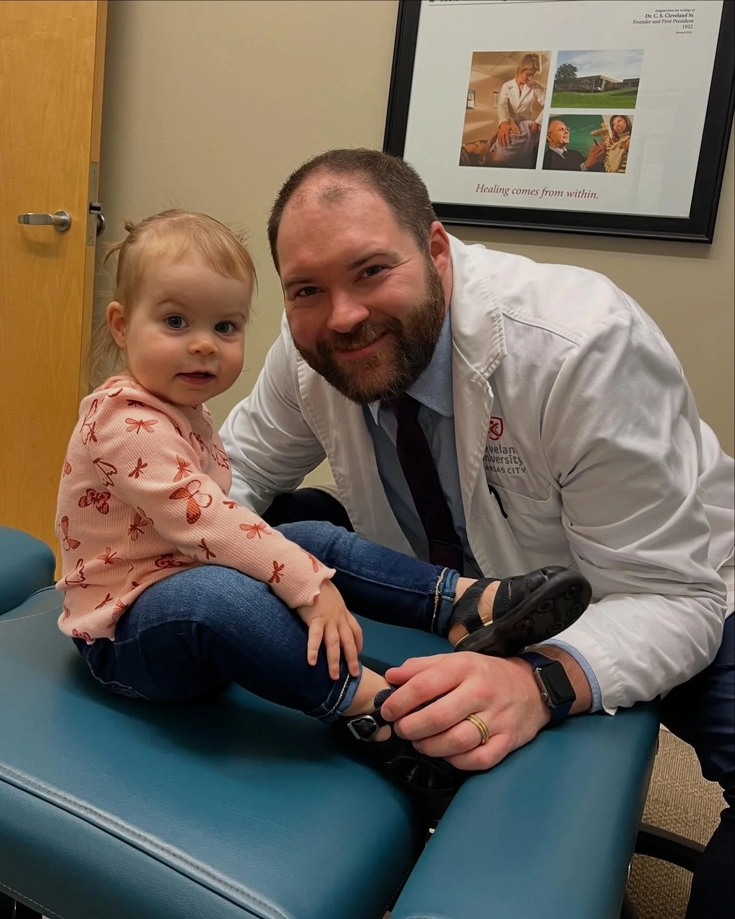 A young girl sitting on an examination table with a male healthcare professional, likely a doctor, who is smiling at the camera. The girl has light red hair, big blue eyes, and is wearing a pink sweatshirt with butterfly patterns. The healthcare professional is wearing a white coat and a wristwatch, and has a beard and short hair. There is a framed poster on the wall behind them with images of medical professionals and a quote that reads, "Healing comes from within."