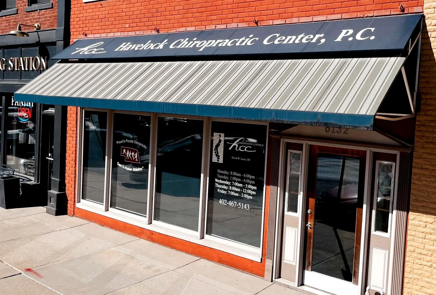 Exterior view of Havelock Chiropractic Center P.C. storefront with a striped awning, glass door, and windows displaying clinic hours and contact information. Adjacent building has a red brick exterior.