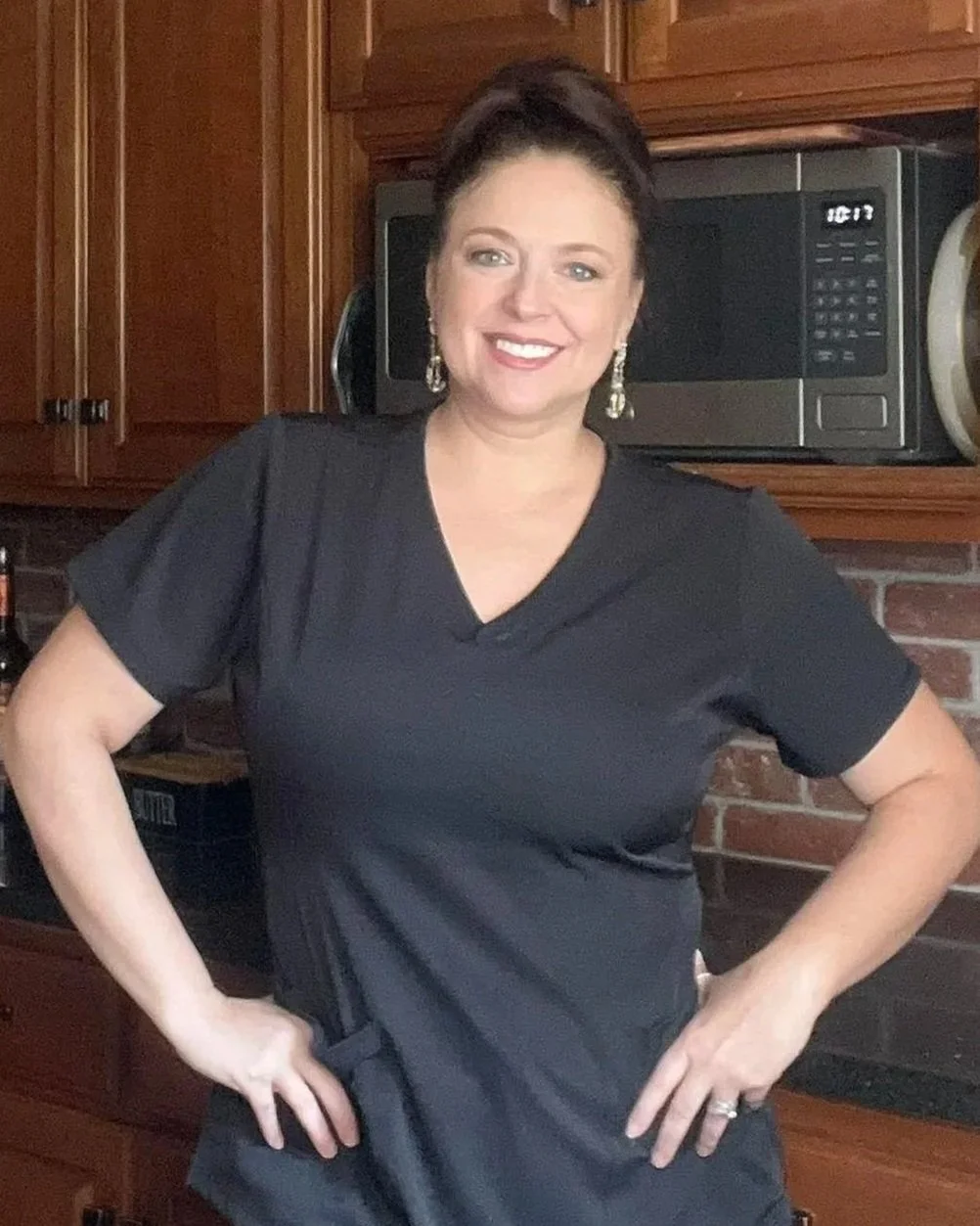 A woman with dark hair in an updo, wearing earrings and a black V-neck top, standing in a kitchen with wooden cabinets and a microwave behind her, smiling with hands on her hips.