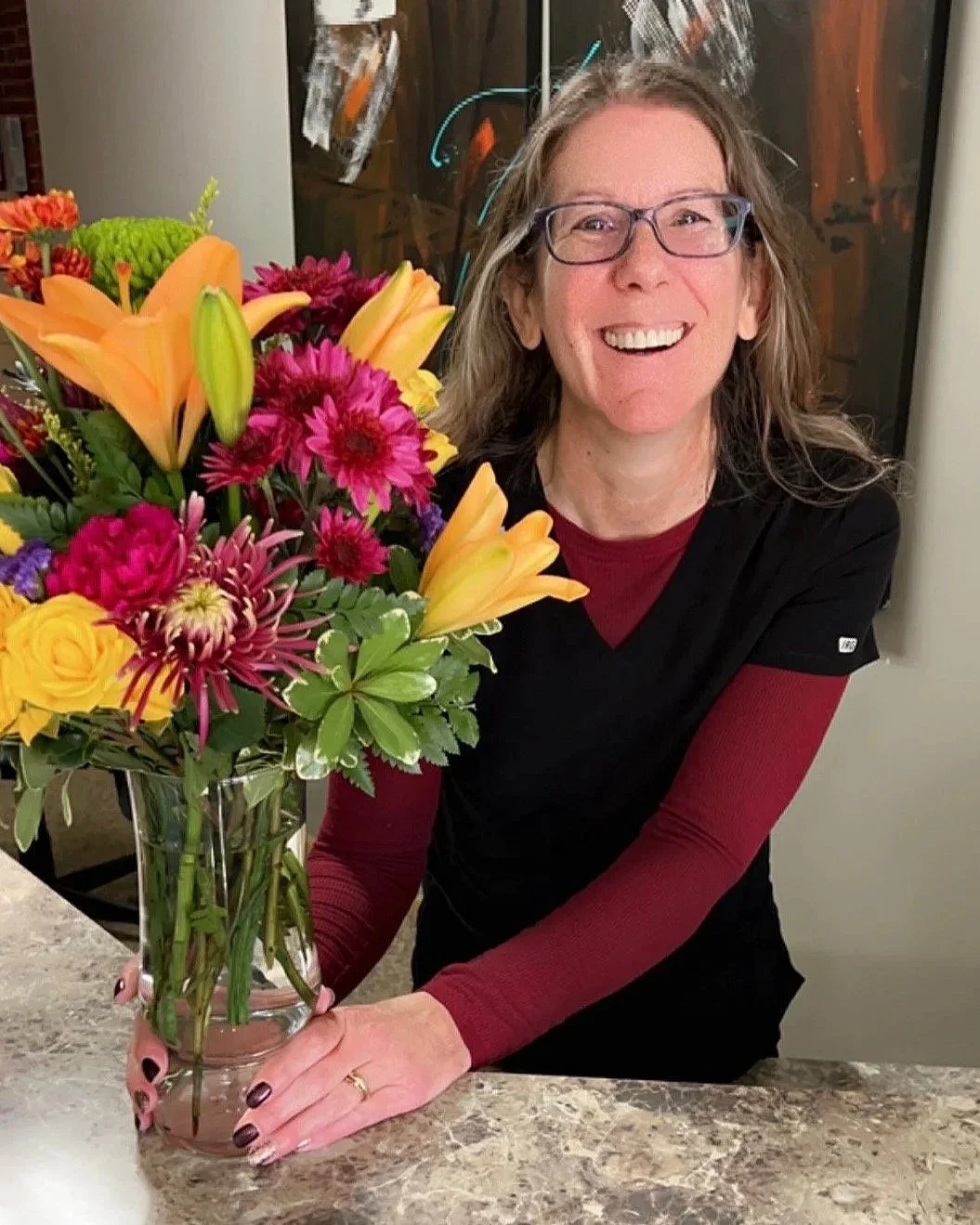 A woman with glasses and long hair smiling while holding a large colorful bouquet of flowers in a glass vase on a marble countertop.