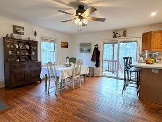 Dining area with a wooden dining table and chairs, a hutch displaying dishes, a ceiling fan, and sliding glass doors leading to an outdoor balcony with a view of trees.