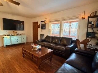 Living room with a brown leather sofa, a matching armchair, a light blue sideboard, a wooden coffee table with books, and a black flat-screen TV mounted on the wall. Large windows with white blinds let in natural light.