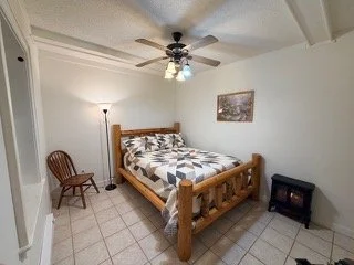 Bedroom with a wooden bed, patterned quilt, wooden chair, floor lamp, ceiling fan, and small black nightstand.