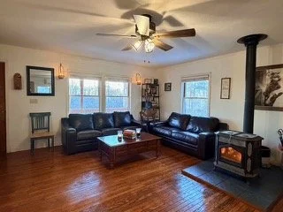 Living room with two black leather sofas, a wooden coffee table, a wood stove, a ceiling fan, large windows, and various wall decorations.