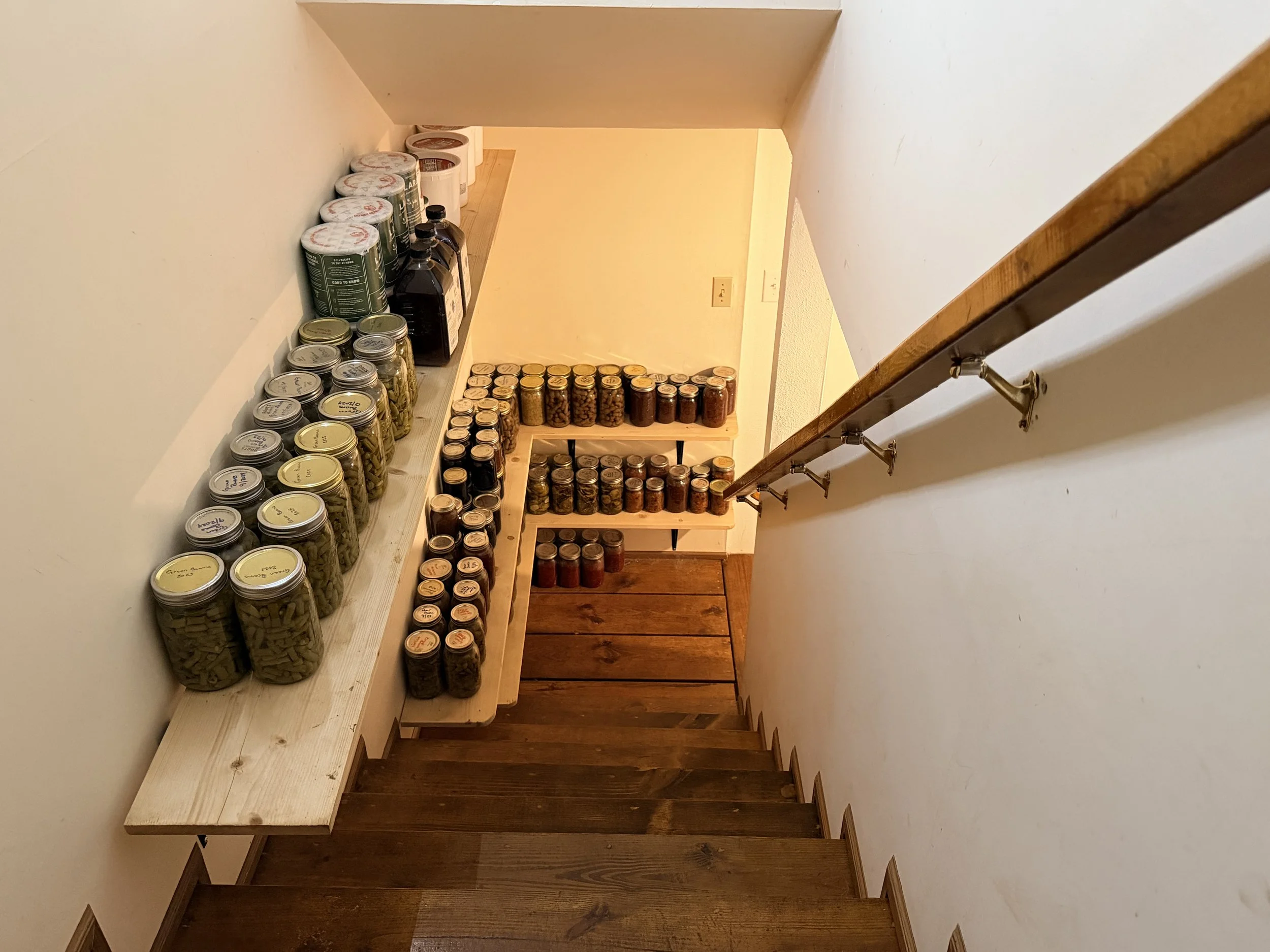 Staircase with wooden steps and a handrail, leading down to a small landing with shelves holding jars of food and canned goods.