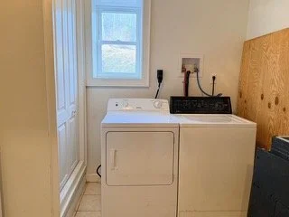 A laundry room with a white top-loading washing machine and a matching dryer, a window above them, and a wooden board leaning against the wall.