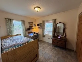 A bedroom with a wooden bed, a dresser with a mirror, two windows with curtains, a desk with a chair, and various framed pictures on the wall.
